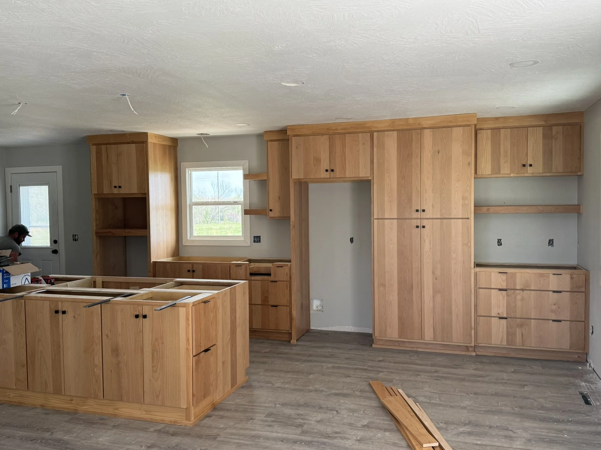 Interior of a kitchen under construction with wooden cabinets, some open spaces, and a window showing outdoor scenery.