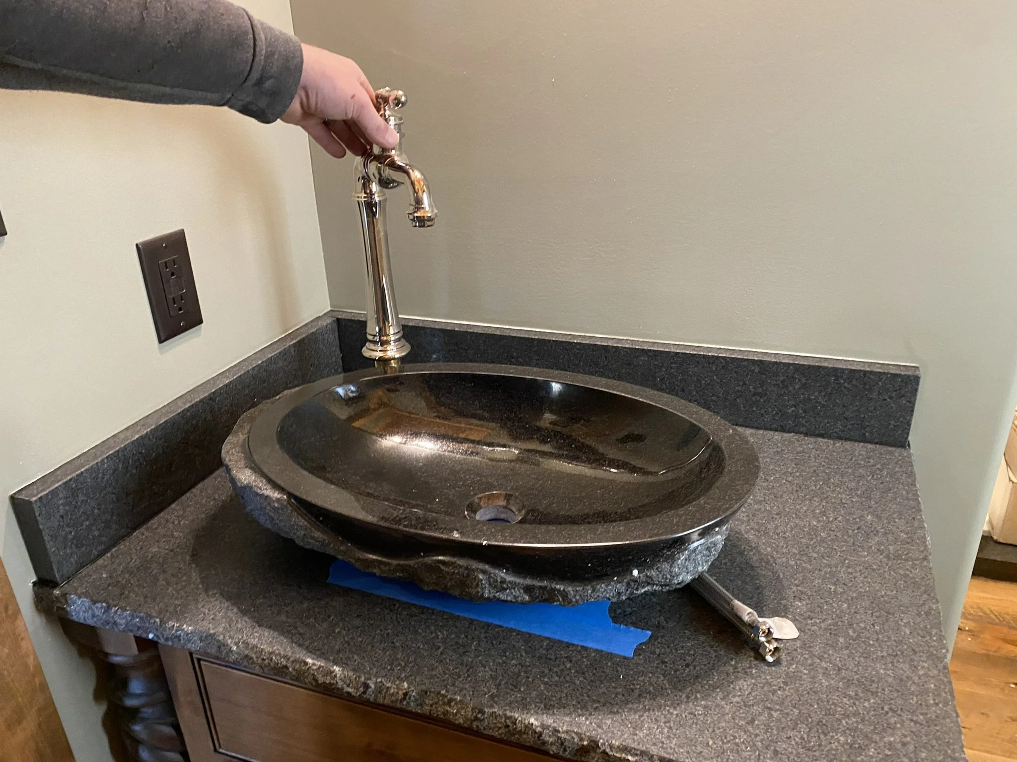 A dark granite kitchen sink with a vintage-style brass faucet, installed in a black granite countertop with a beige wall behind it.