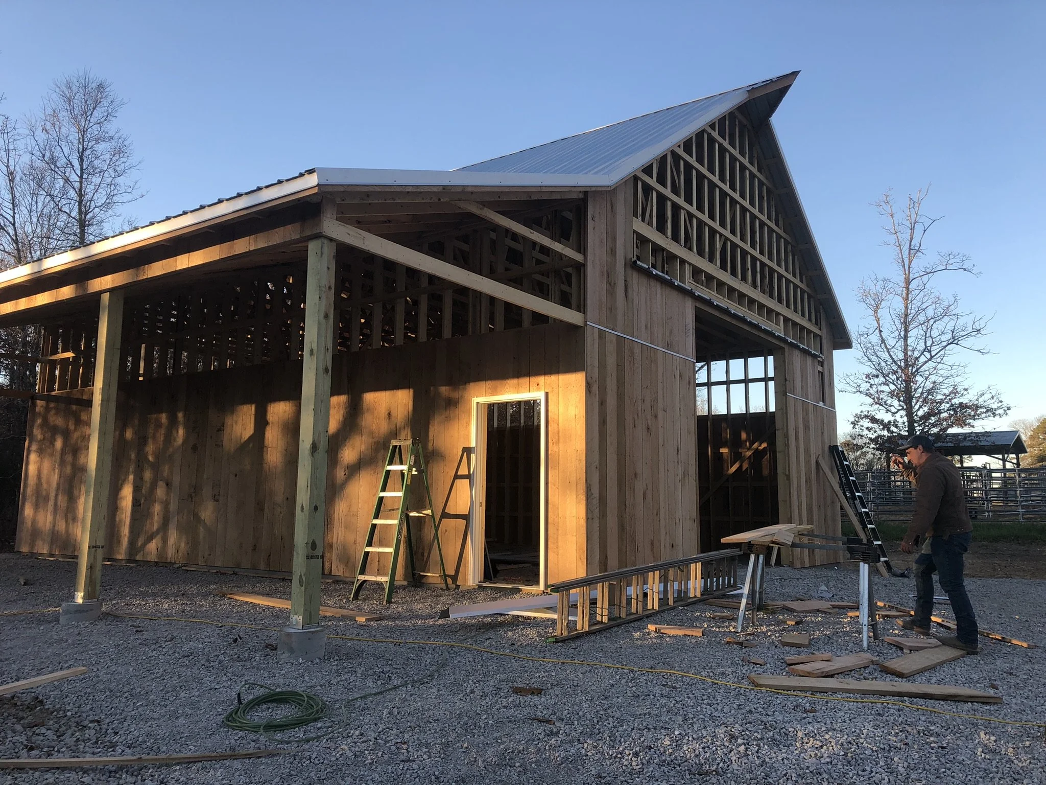 A two-story wooden building under construction with an angled metal roof, surrounded by trees and construction tools, with workers present.