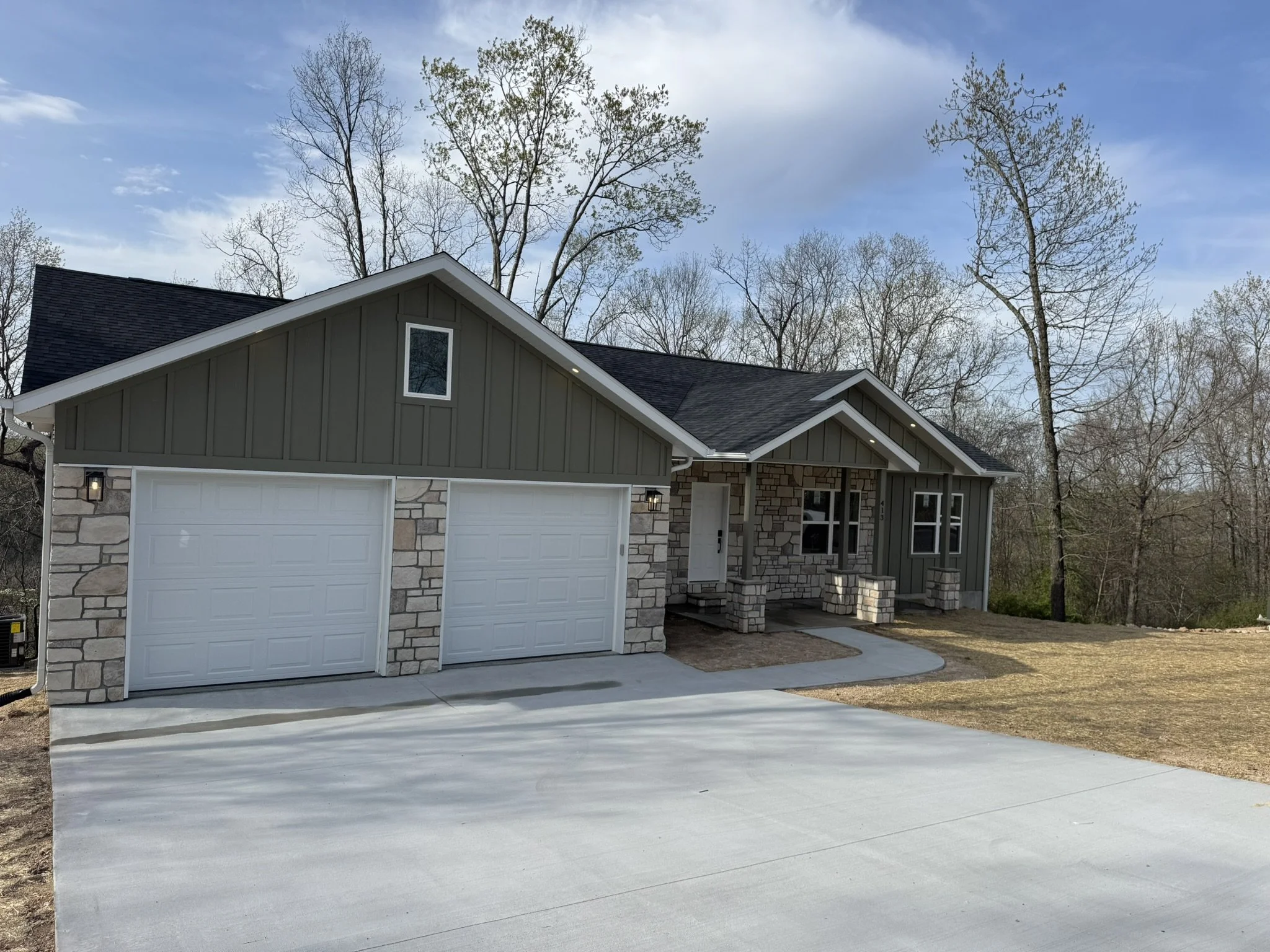 A house with a gray exterior, stone accents, and a dark roof, featuring a white garage door, with trees in the background and a bright blue sky.