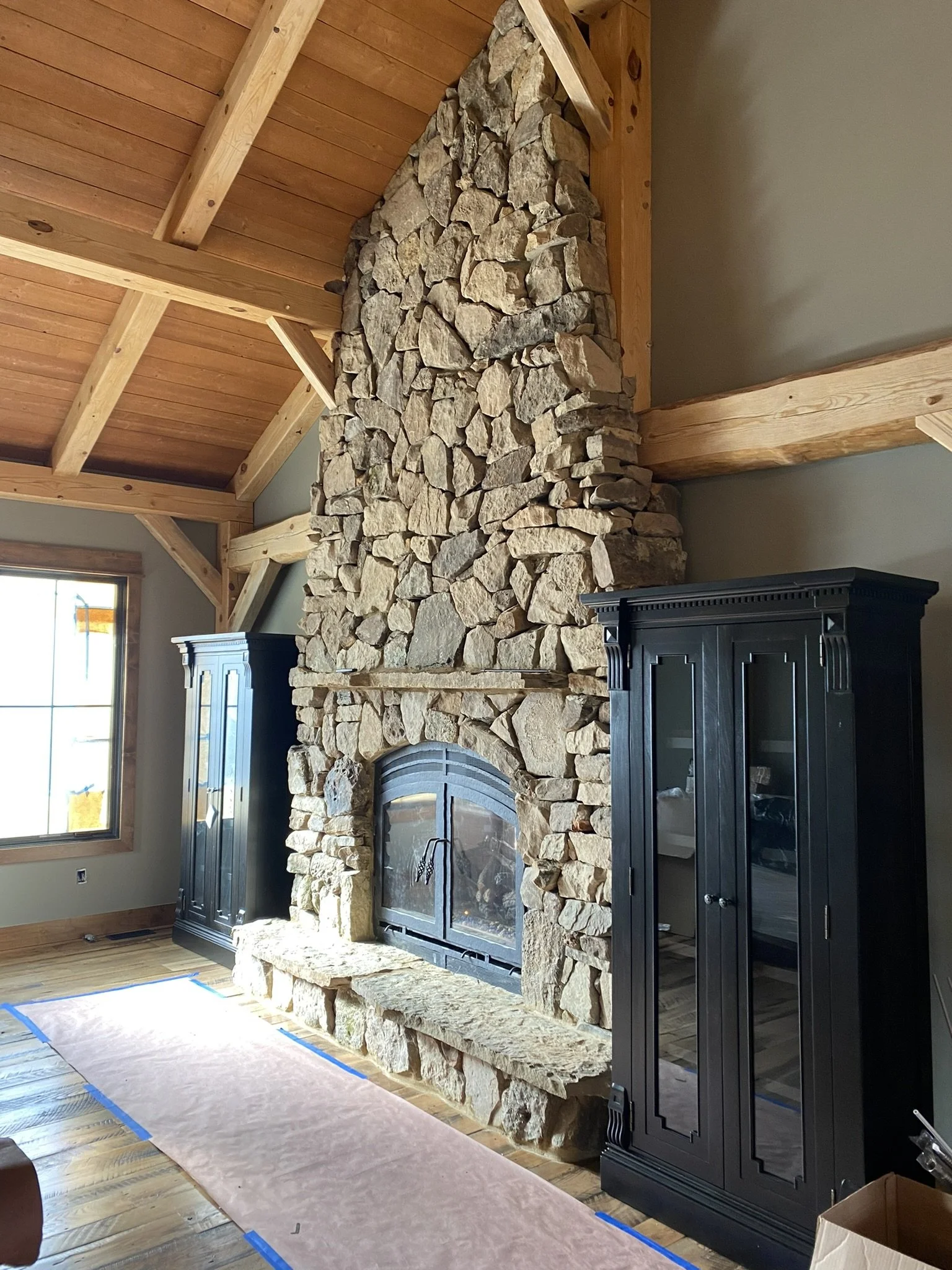 A tall stone fireplace in a living room with a wooden ceiling and a window to the left. There is a black cabinet in front of the fireplace and a paper covering the wooden floor.