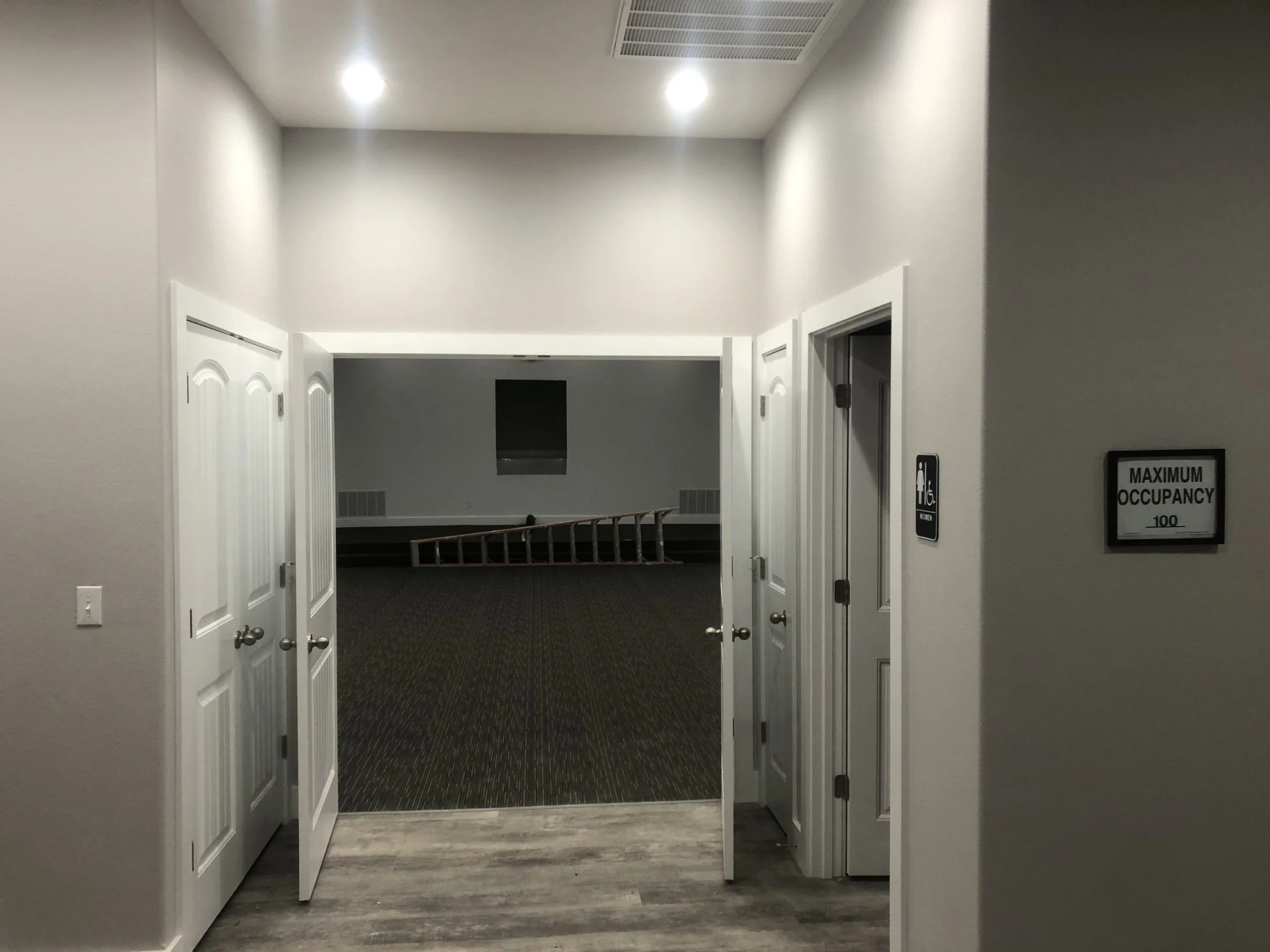 A hallway with open double doors on each side, leading into rooms, with a row of chairs at the far end and a sign indicating maximum occupancy.