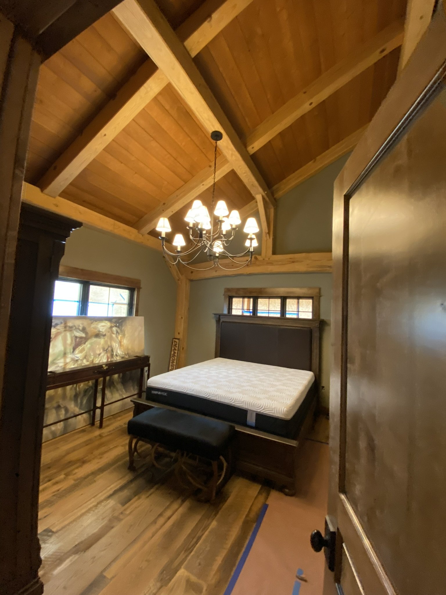 A bedroom with wooden ceilings and walls, a chandelier with multiple lights hanging from the ceiling, a bed with a white mattress, a dark wooden headboard, a piano with a colorful painting on top, a window, and a mirror on the right wall.