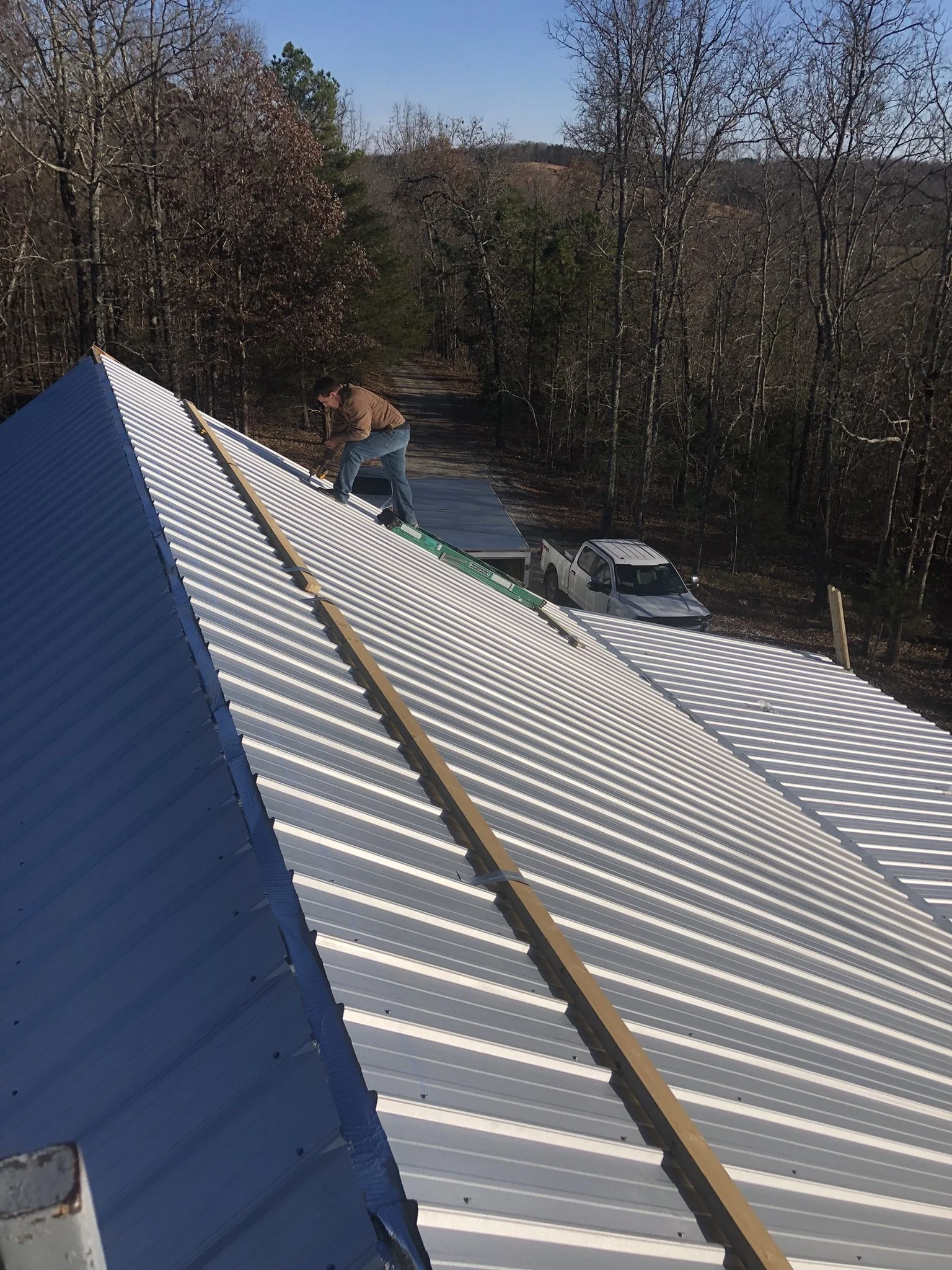 A man installing or repairing a silver metal roof on a building, with a truck parked nearby and a wooded area in the background.