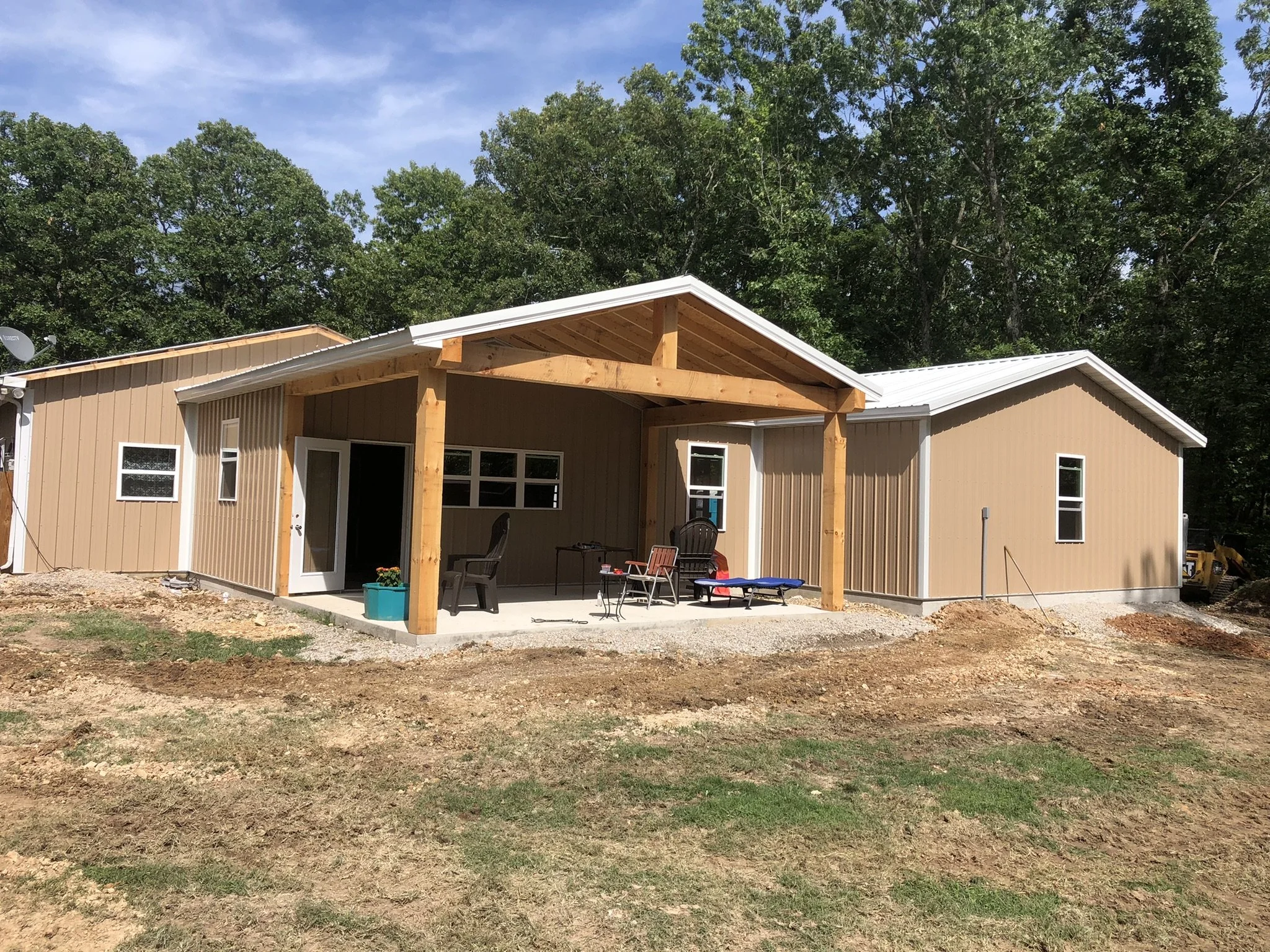 A house under construction with beige siding, a white metal roof, and a wooden porch frame. There are outdoor chairs and a small table in the backyard area, with trees in the background and a blue sky.