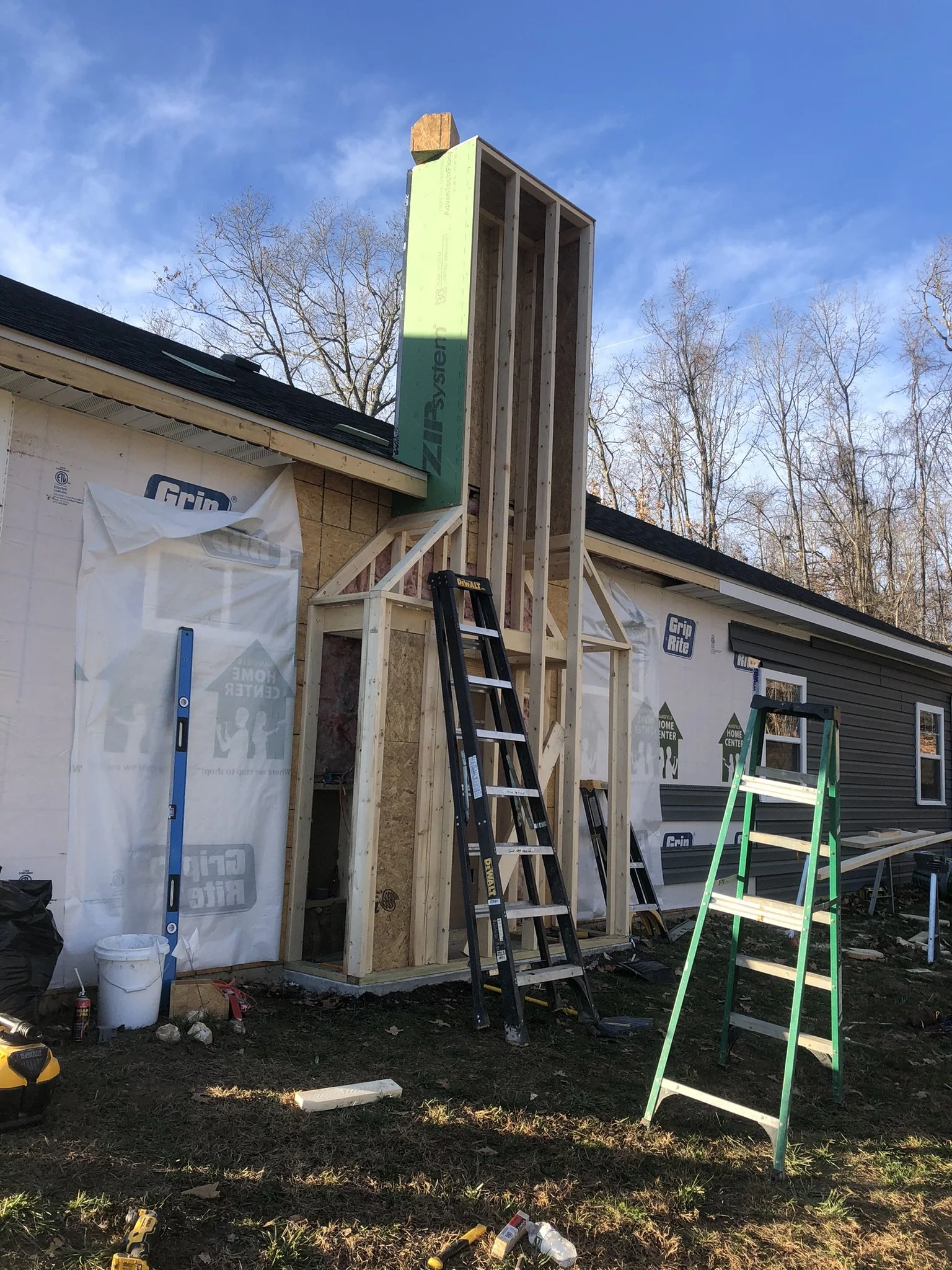 Construction site with a partially built house extension, including framing and sheathing, ladders, and tools on a grassy ground, under blue sky with trees in background.