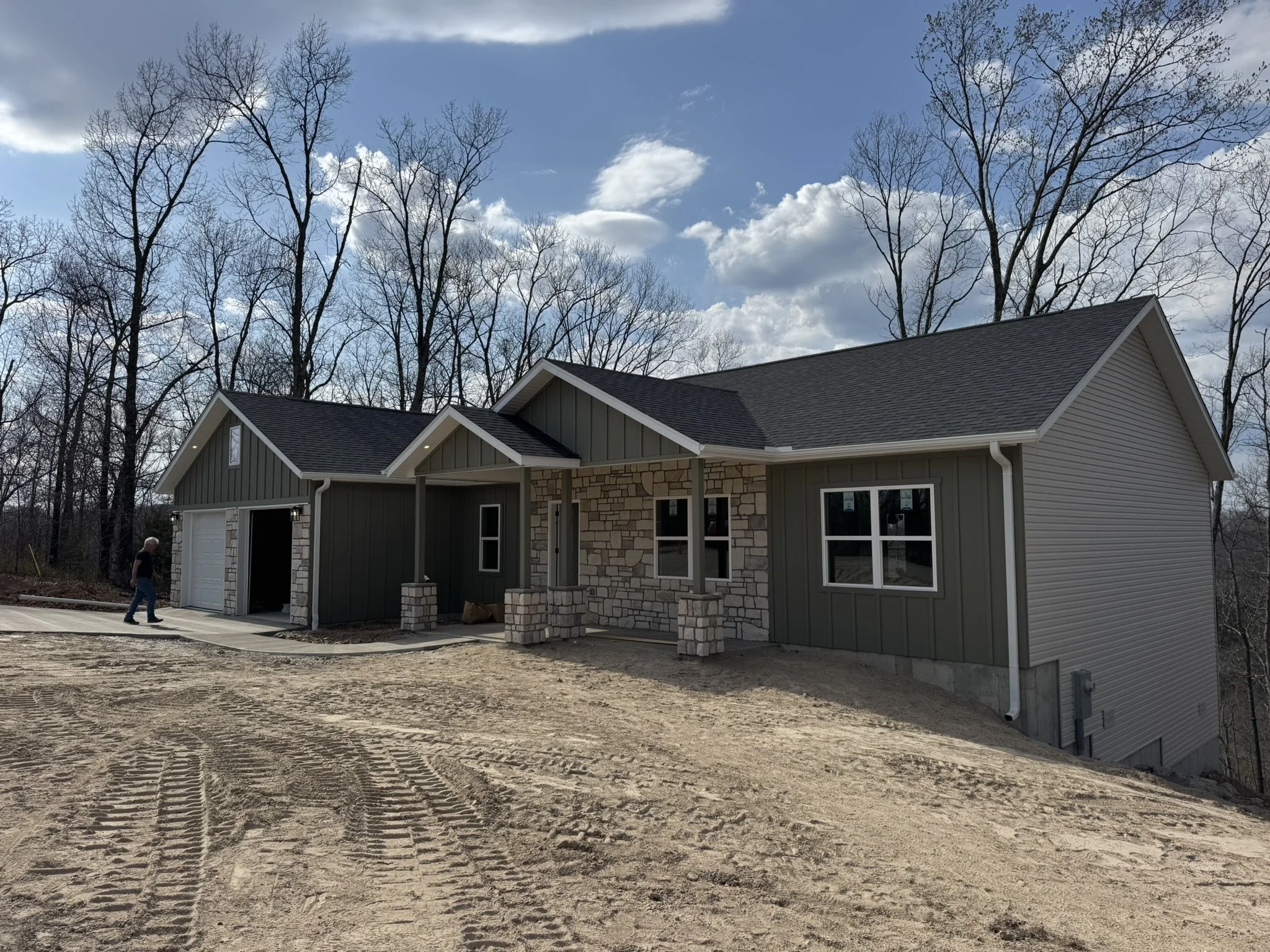 A house under construction with a paved driveway, surrounded by leafless trees, with a bright blue sky with some clouds.