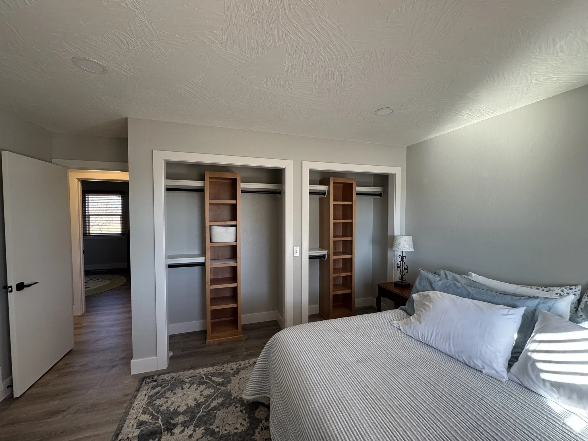A bedroom with a bed covered in blue striped bedding, two pillows, and a decorative pillow. There is a small wooden nightstand with a lampshade, and built-in open wooden shelving on the wall above the bed. A window with blinds is visible in the backg