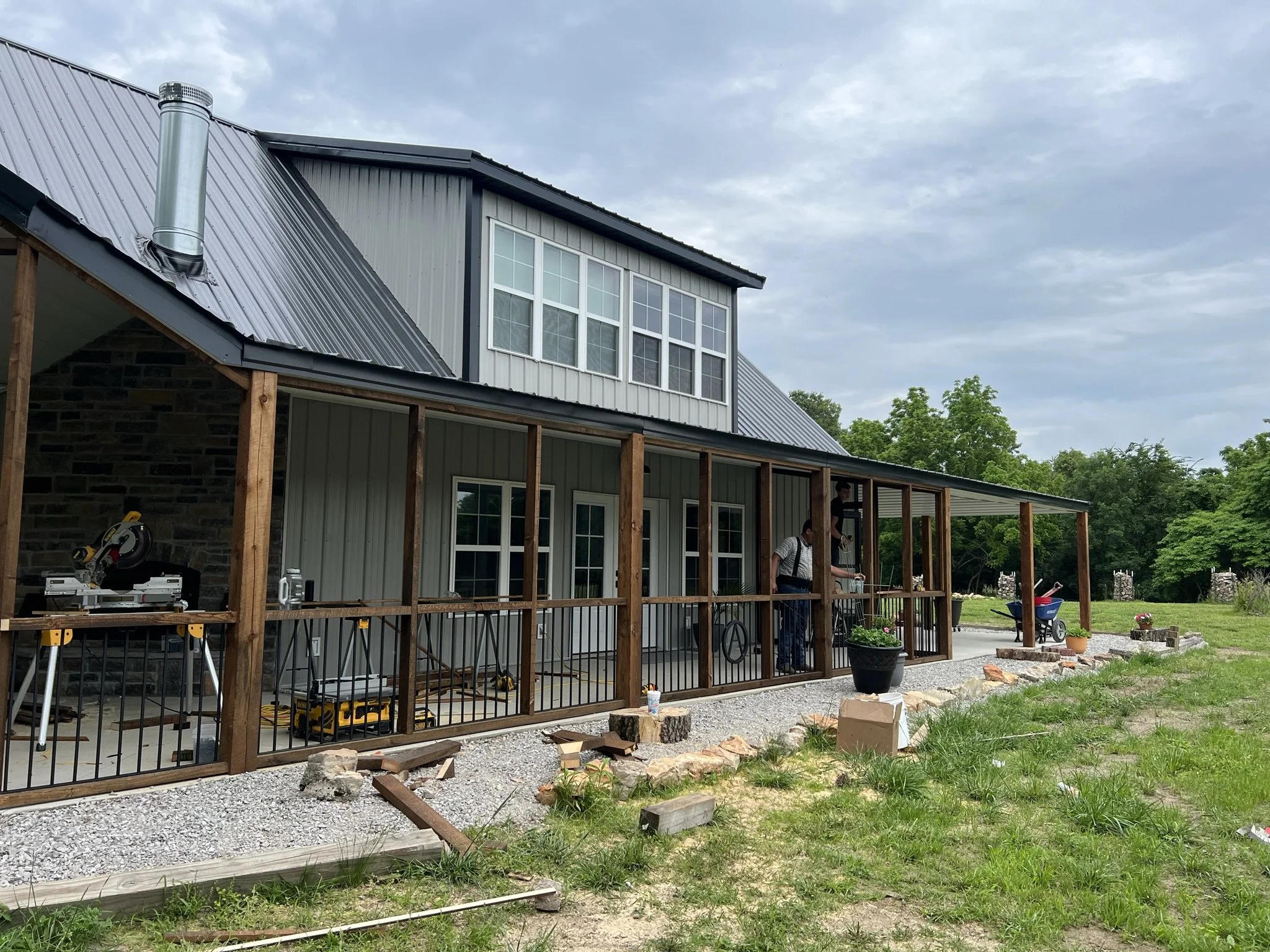 A house under construction with a partially finished porch, wooden support beams, and construction tools outside. The house has gray siding, large windows, and a metal roof, with a cloudy sky in the background and green trees nearby.