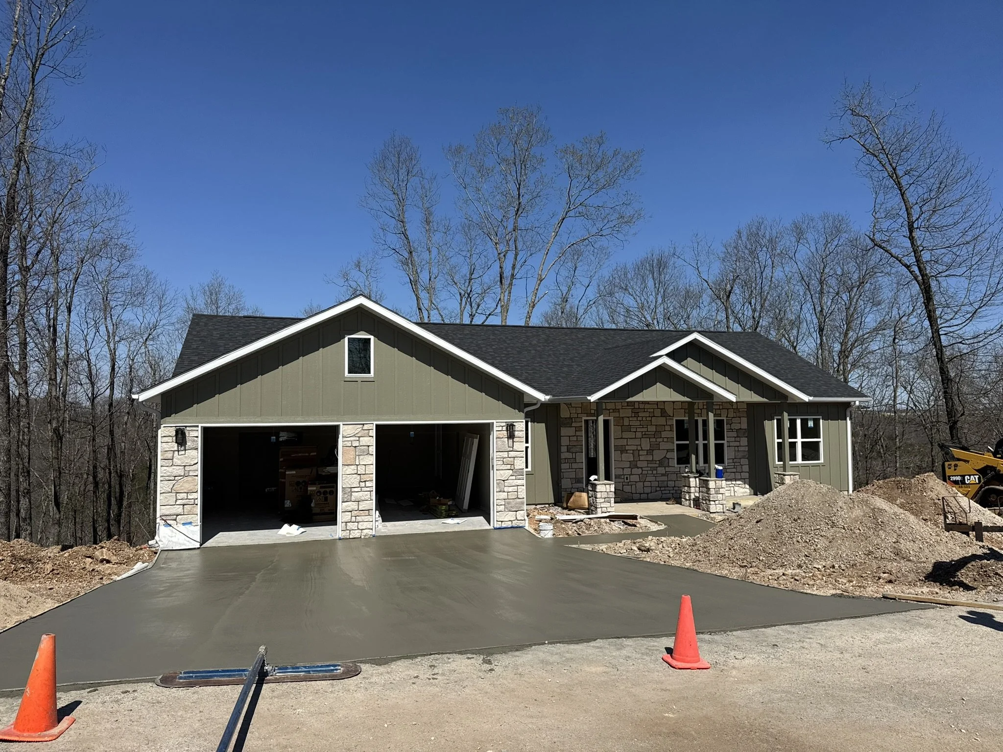 Newly constructed house with a concrete driveway, construction cones, and a garage opening. The house has a stone and siding exterior with a gabled roof. There are construction materials and equipment visible, indicating ongoing work.