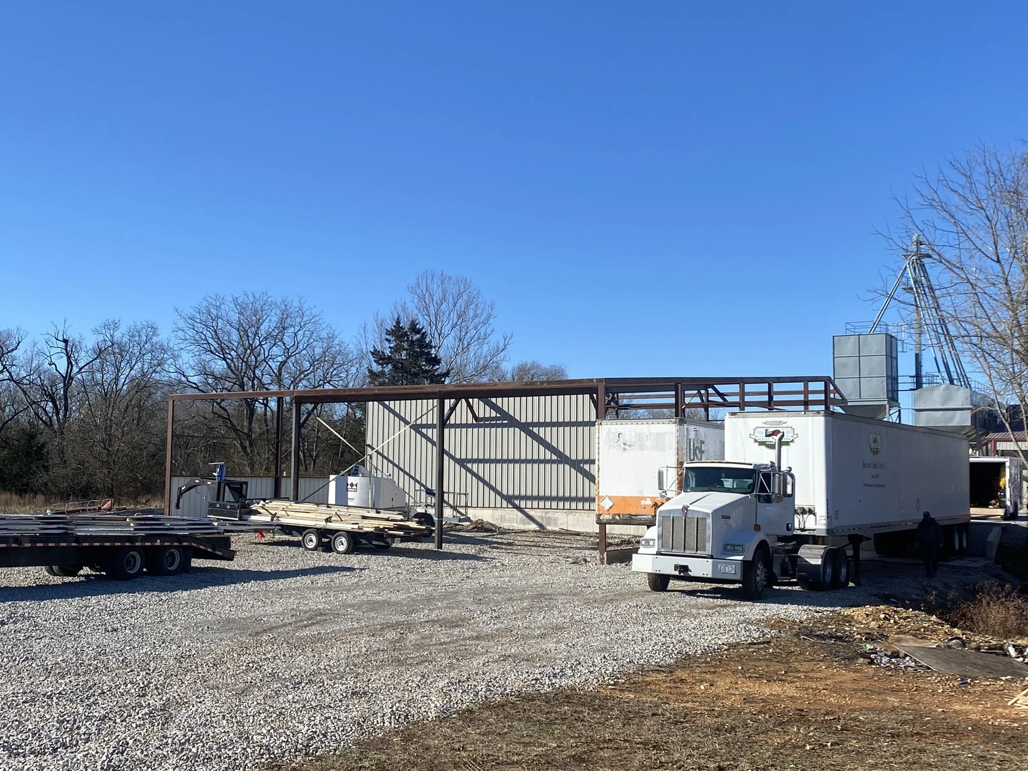 Construction site with building framework, parked trucks, trees, and a clear blue sky.