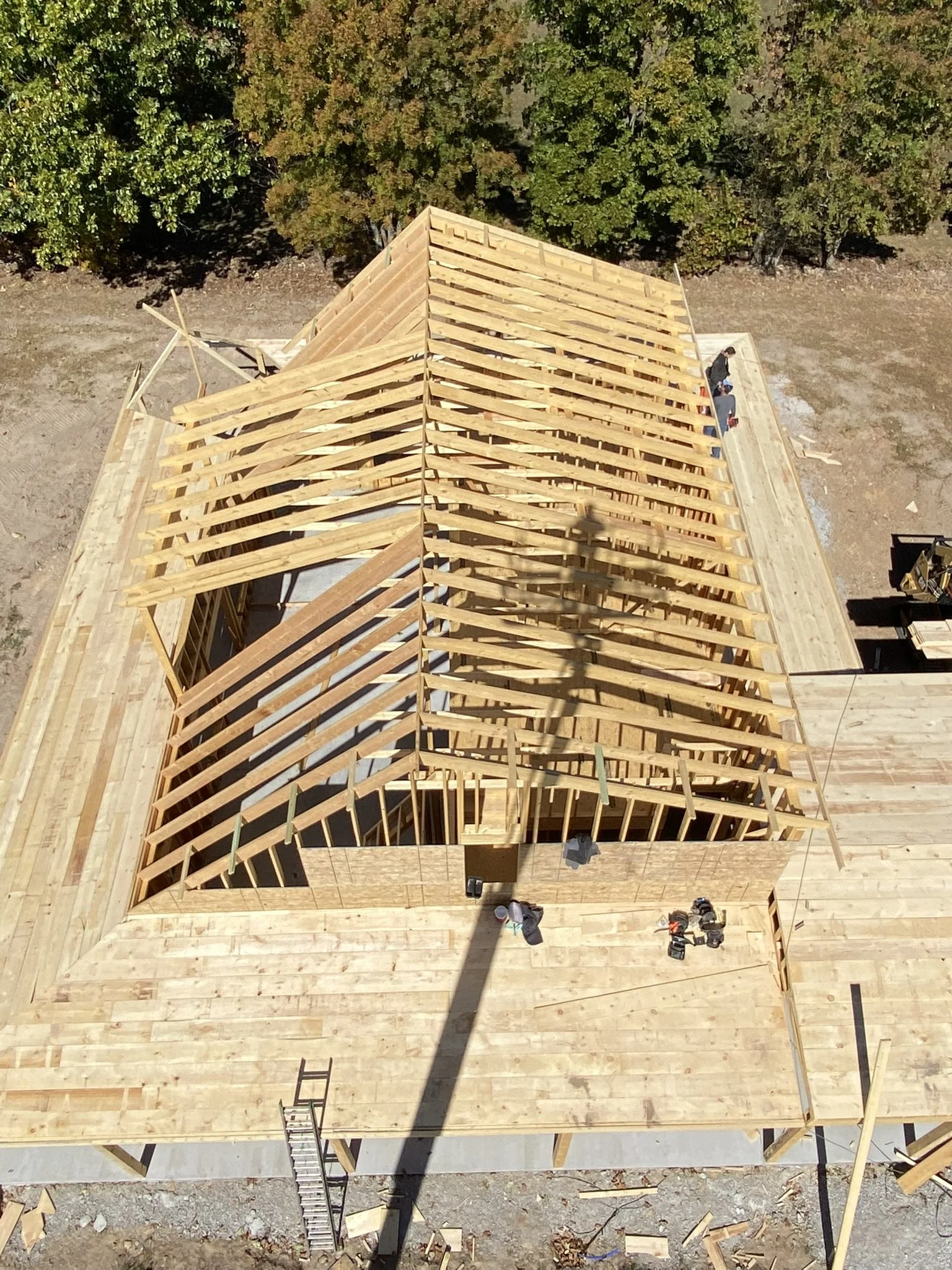 Aerial view of a wooden building under construction, with its roof framework visible, surrounded by trees and construction tools on the ground.