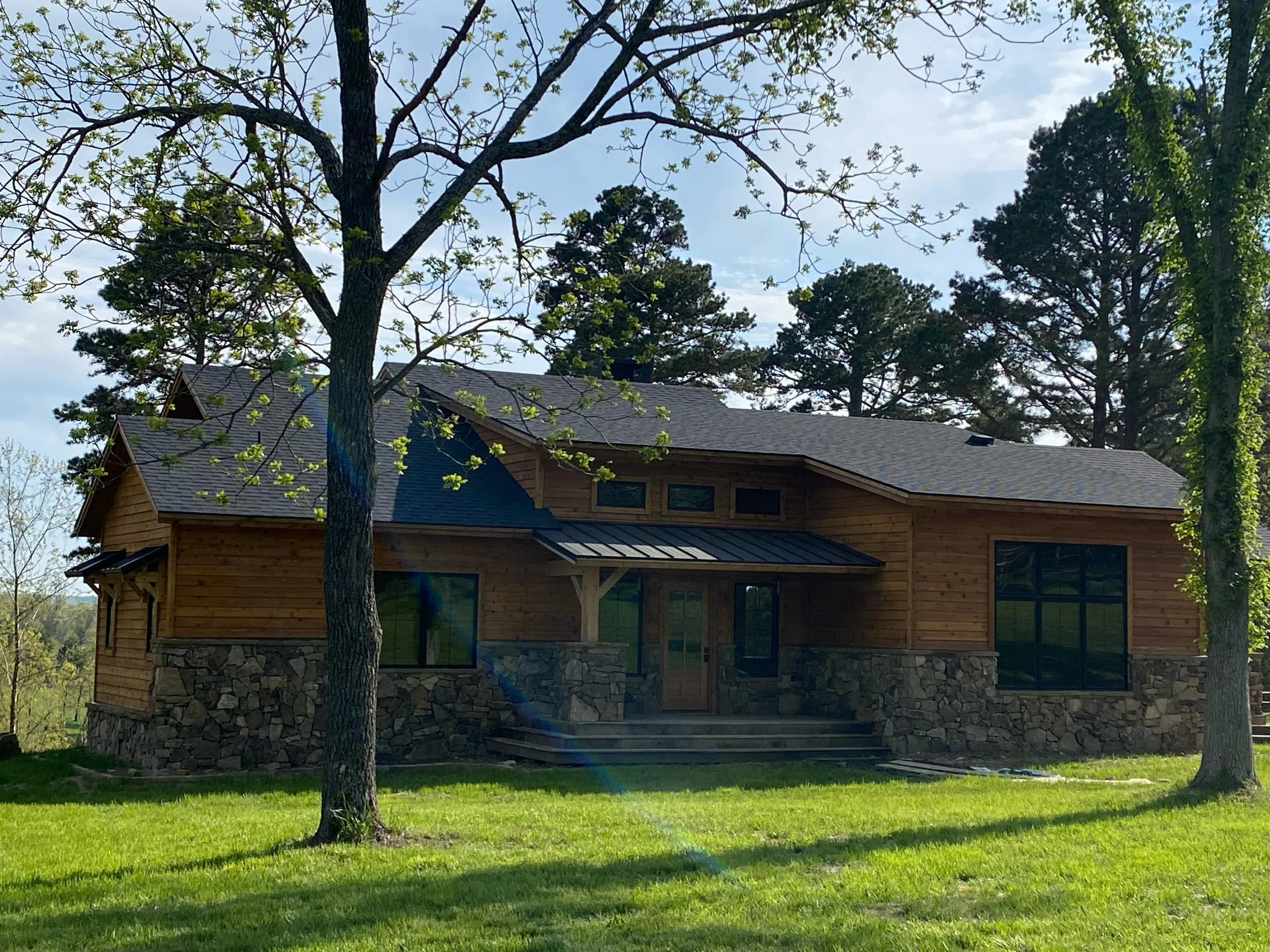 A modern house with dark-colored roof shingles, large windows, and stone accents on the lower part of the exterior walls, surrounded by green grass and trees, under a partly cloudy sky.