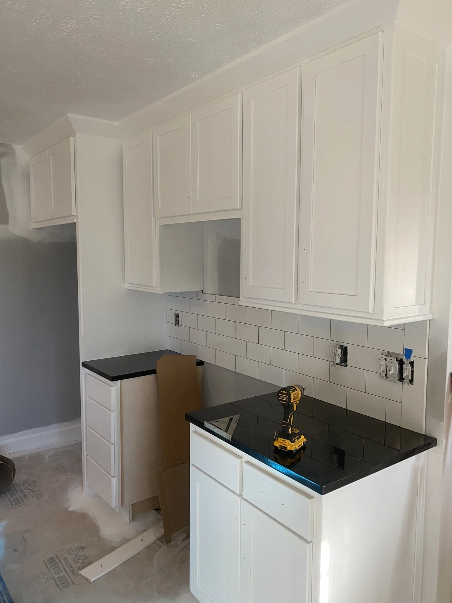 Kitchen cabinets being installed, with tools like a drill on the countertop, white tile backsplash, and unfinished floor.