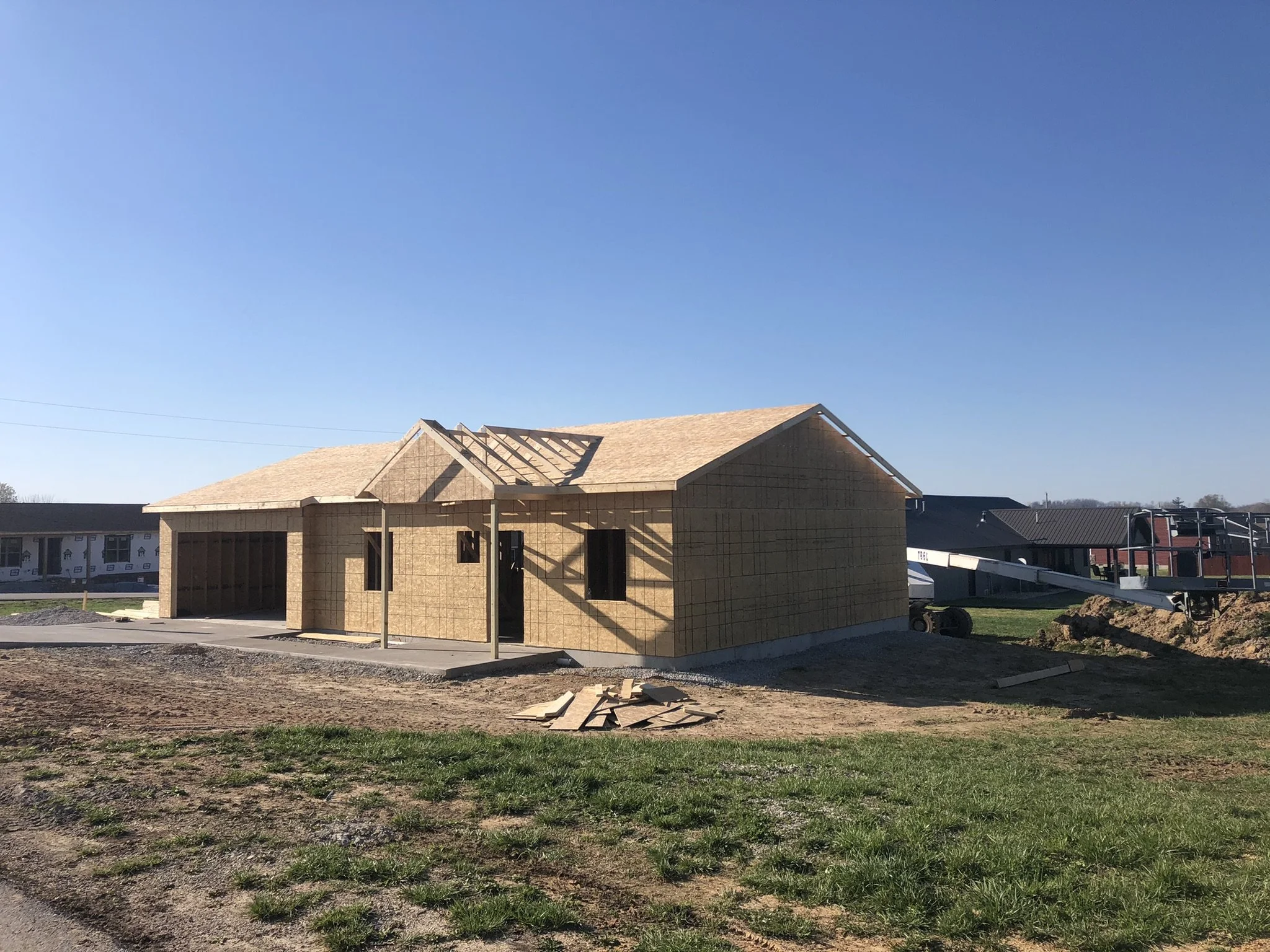 A house under construction with a wooden framework and a roof, scattered construction materials on the ground, and neighboring houses in the background under a clear blue sky.