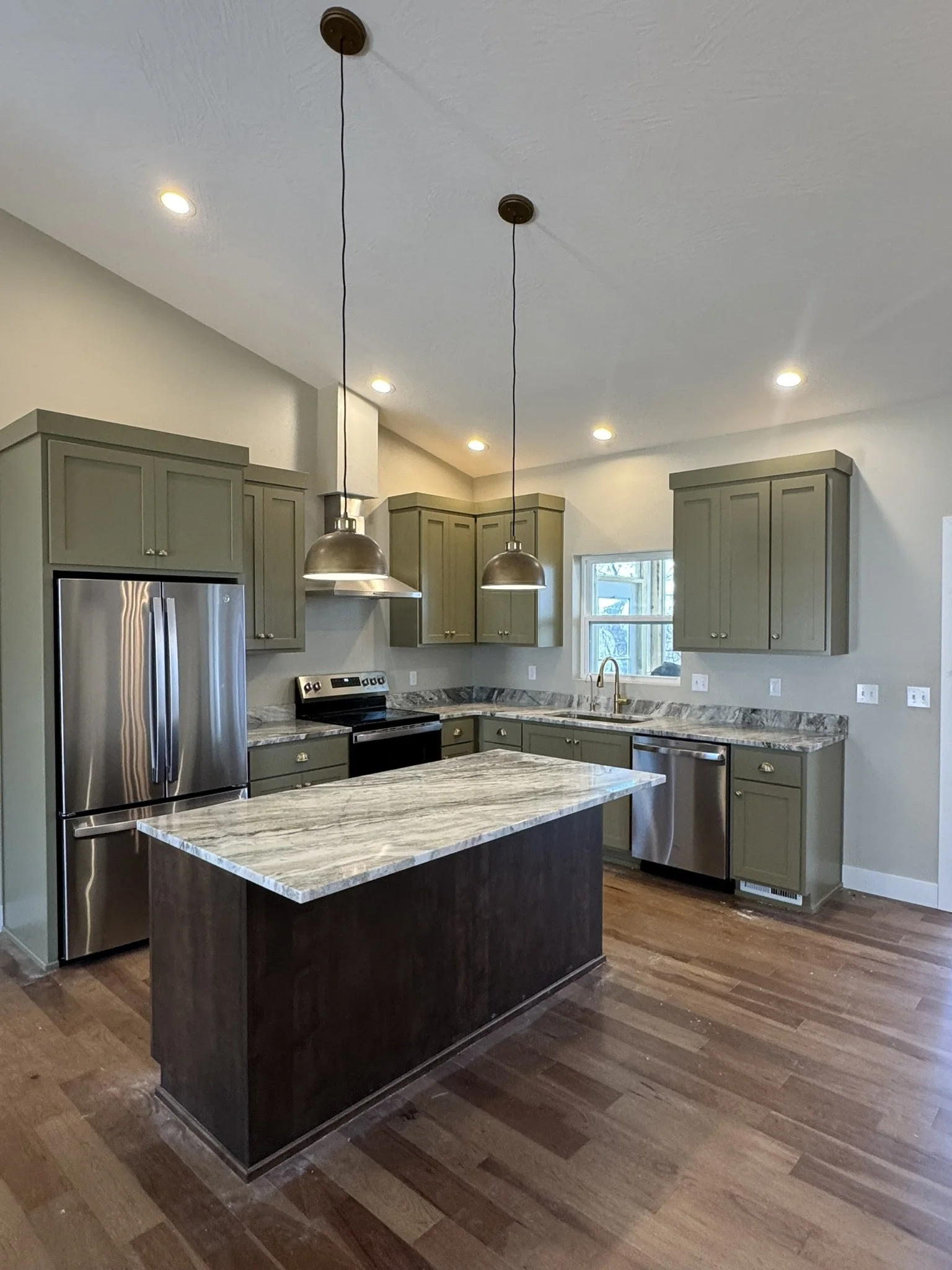 A modern kitchen featuring gray upper cabinets, a stainless steel refrigerator, and a granite countertop island with a dark base. There are two hanging pendant lights above the island, recessed ceiling lights, and a window with a view of the outdoors
