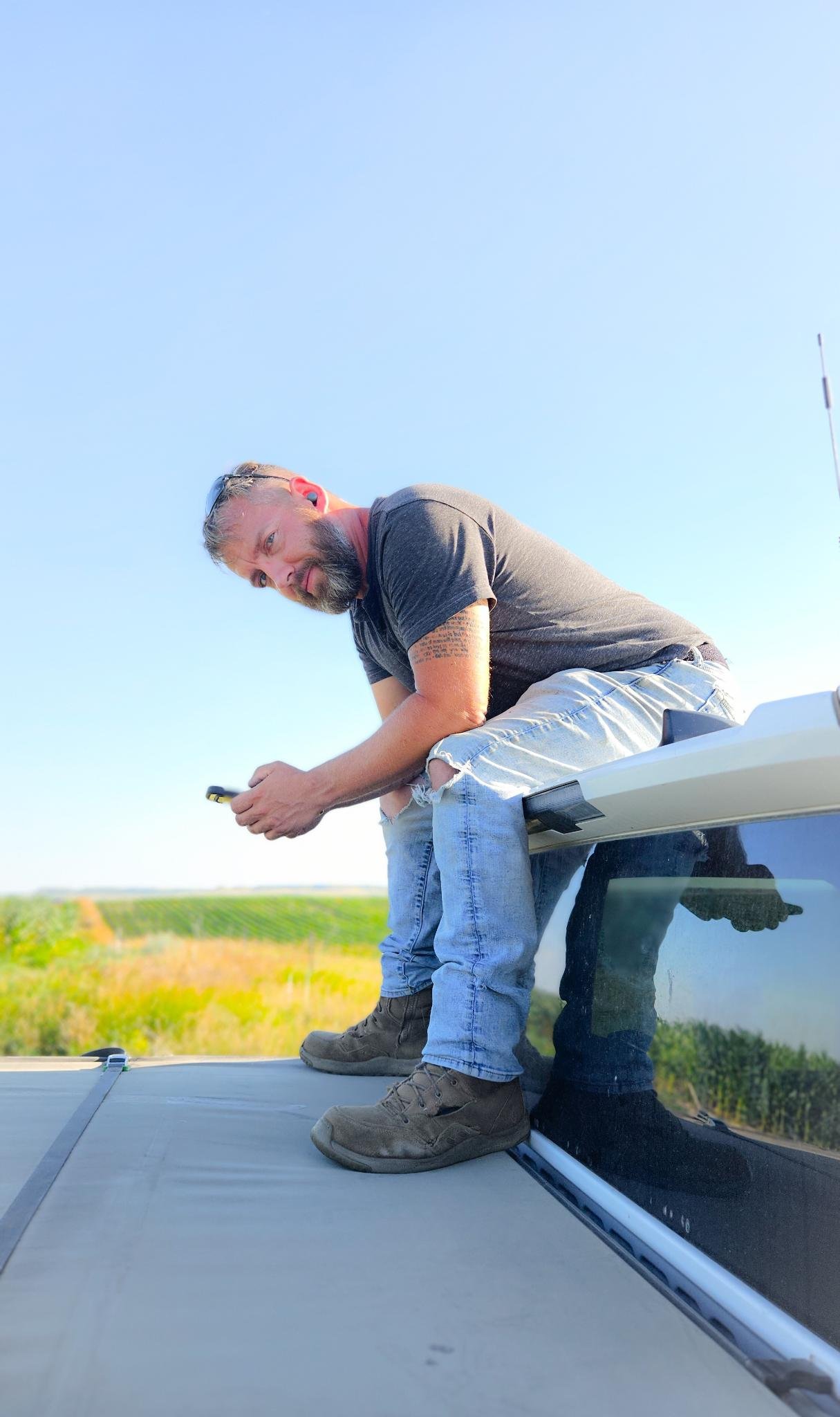 A man with a beard and tattoo sitting on top of a vehicle, holding a phone, in a rural area with green fields under a clear blue sky.