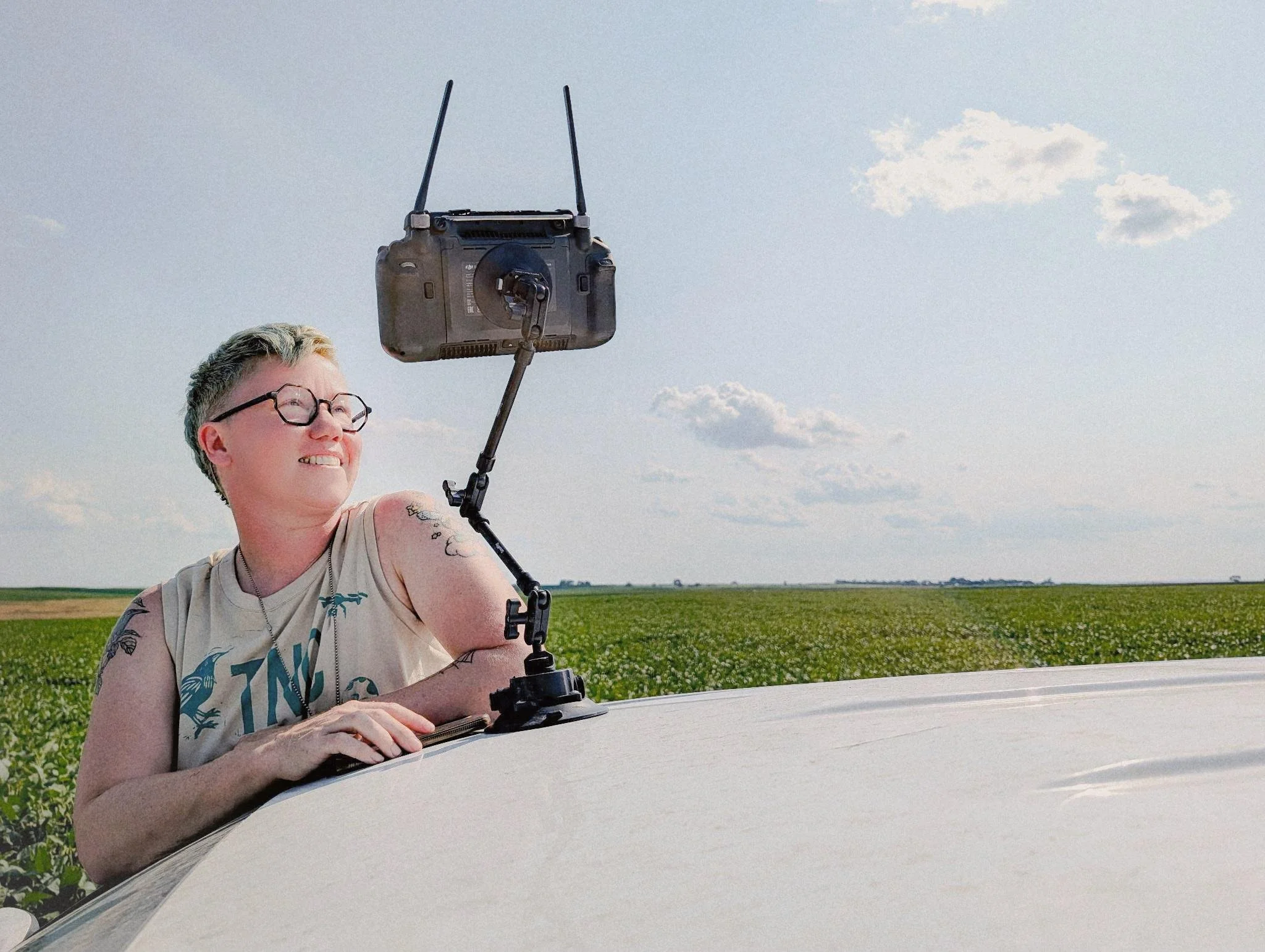Person with short blonde hair and glasses smiling as they hold a remote control in front of a drone during daytime in a rural setting with green fields and a blue sky.