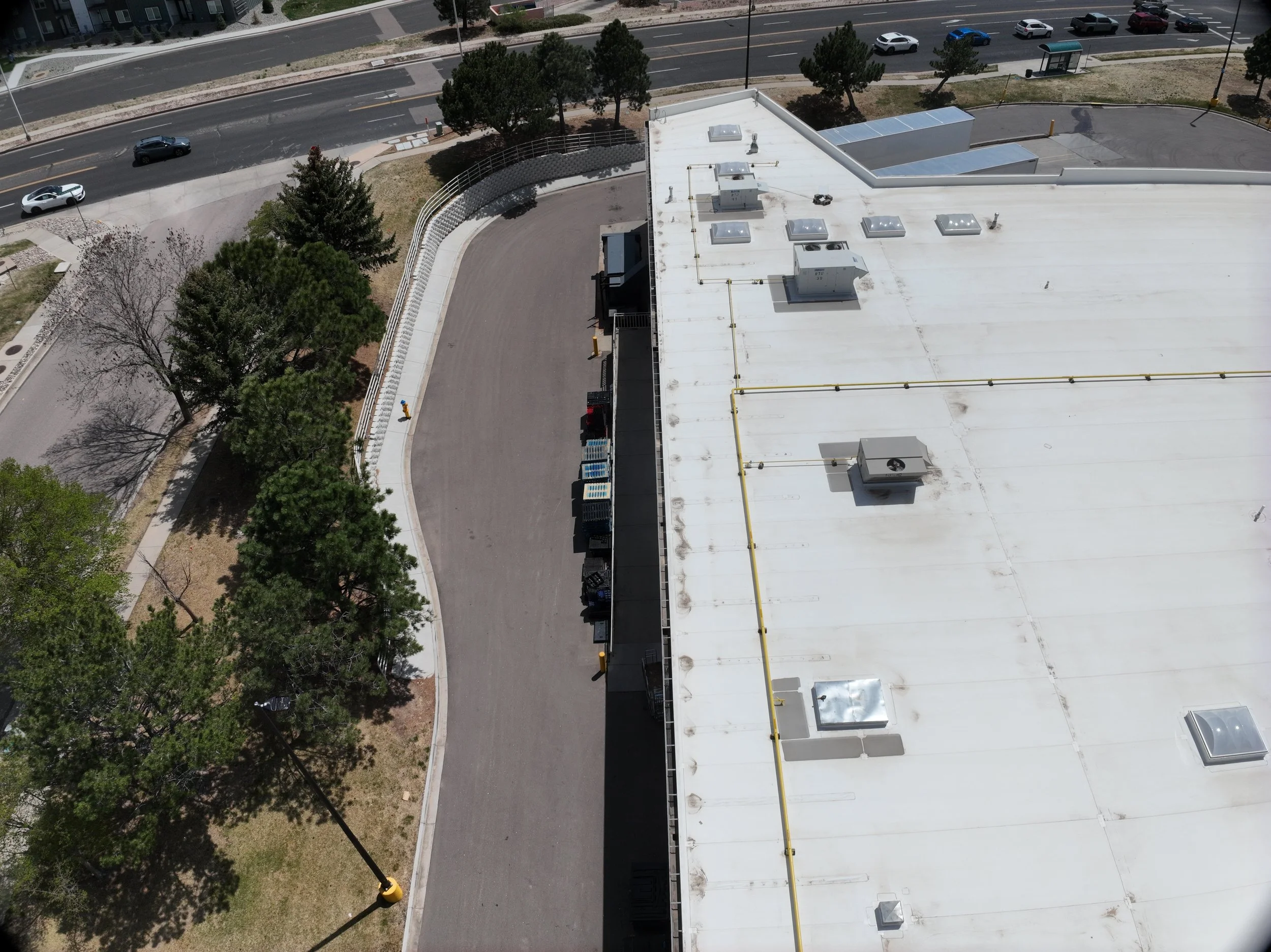 An aerial view of a flat white building with a yellow pipe along the roof, surrounded by trees, a sidewalk, and a parking lot with several cars on a street.