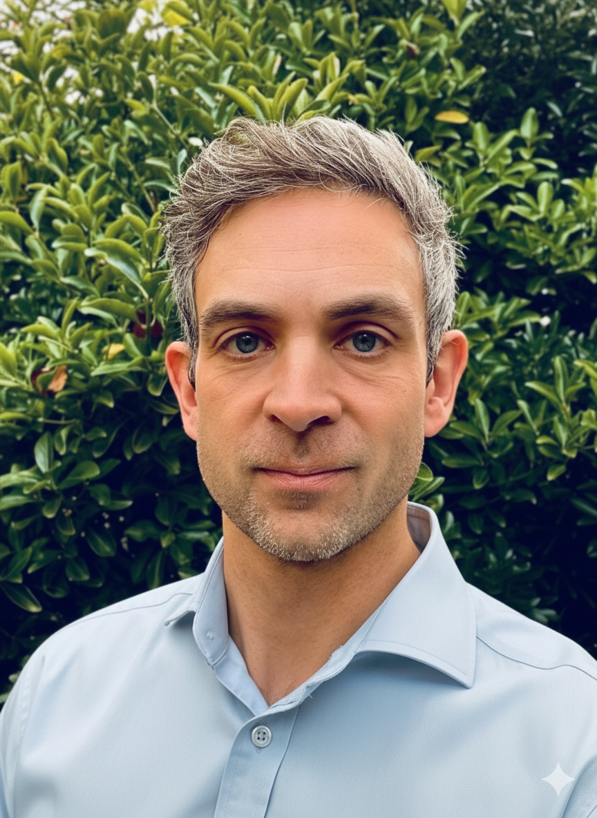A man with gray hair and a beard wearing a light blue collared shirt, standing in front of green bushes.