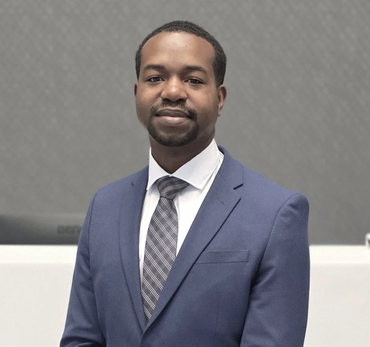 A handsome business consultant in a navy suit, white shirt, and patterned tie standing against a gray background.