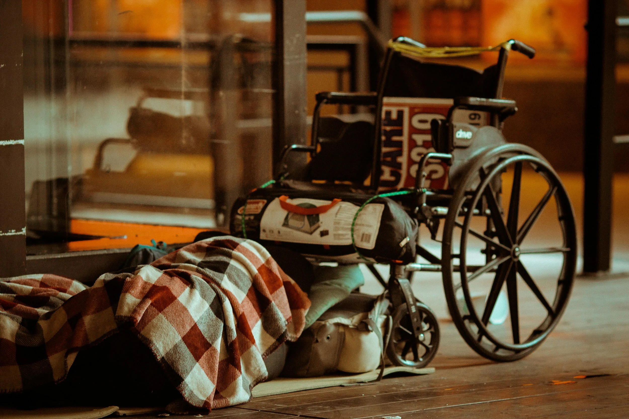 A wheelchair next to a pile of folded clothes or blankets, with various bags and items nearby, on a wooden floor in a room with warm lighting.