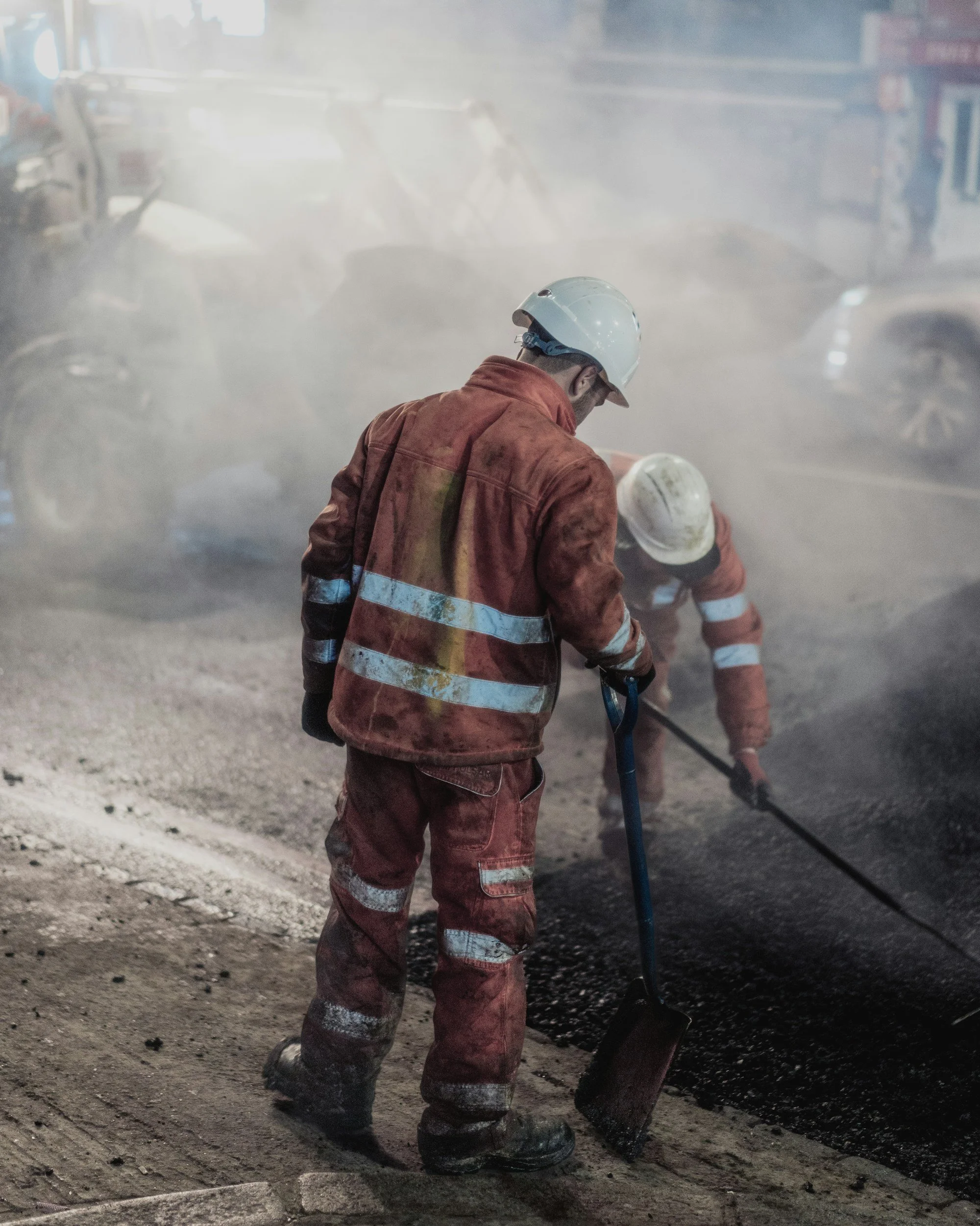 Construction workers wearing orange safety gear and helmets working on paved road in a dusty environment.