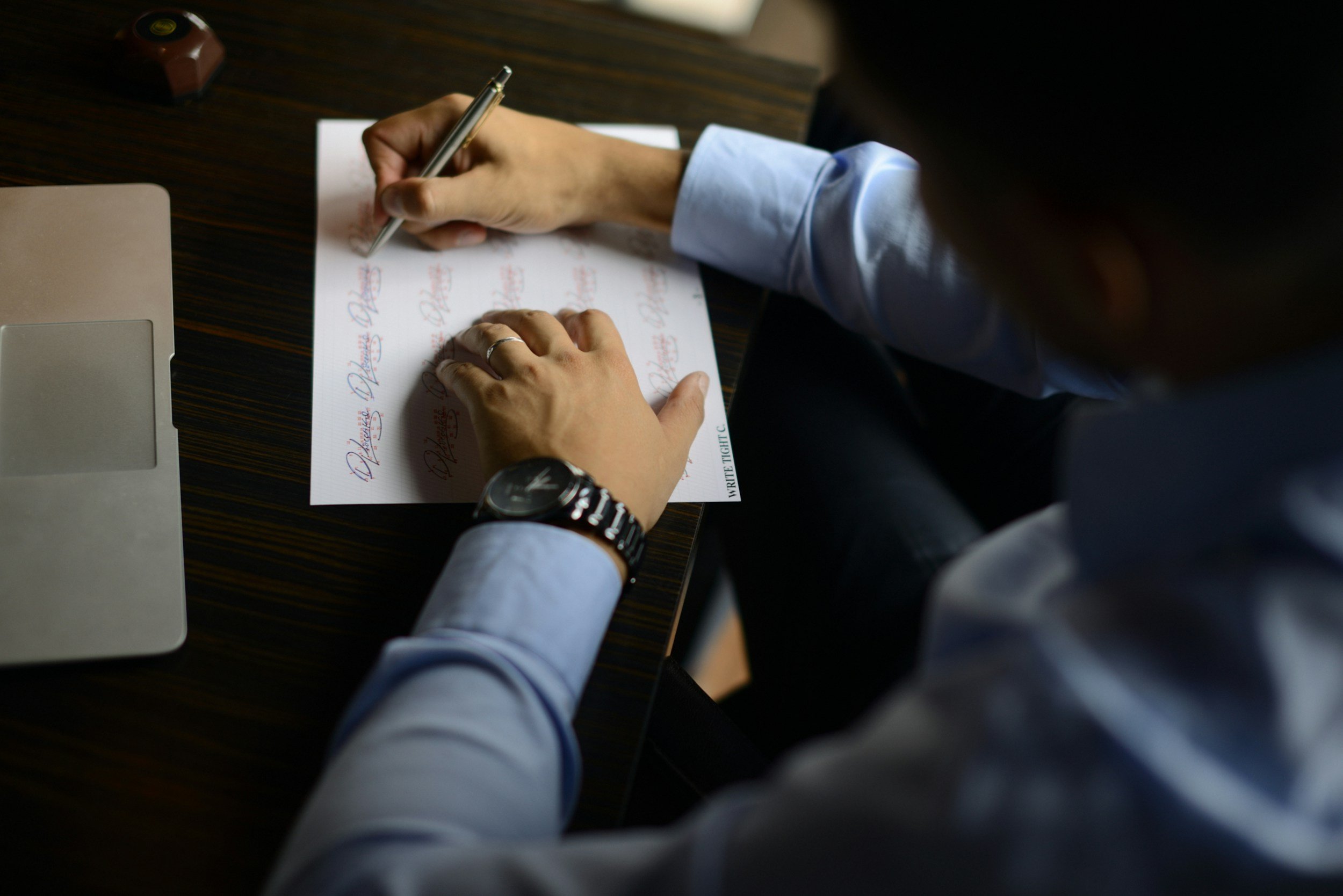 Person writing on a sheet of paper with printed signatures, sitting at a dark wooden desk with a laptop and a stamp in the background.