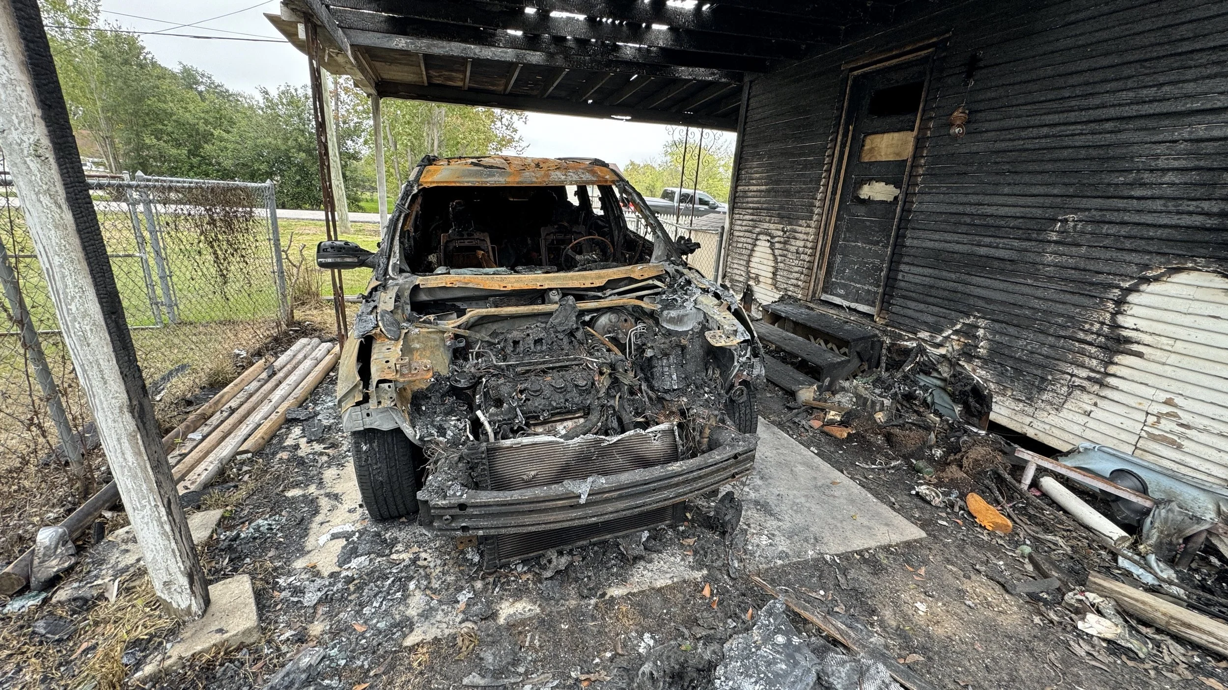 Remains of a burned car with a destroyed engine, parked inside a blackened garage with burnt walls and debris on the ground.
