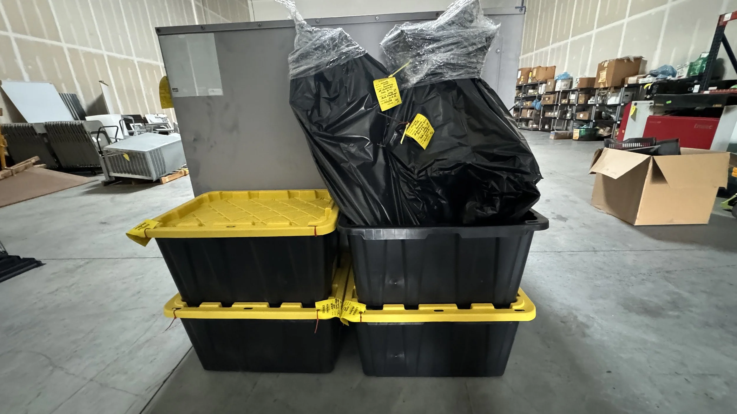 Four large black storage bins, three with yellow lids and one behind with a black trash bag on top, are stacked in a warehouse.
