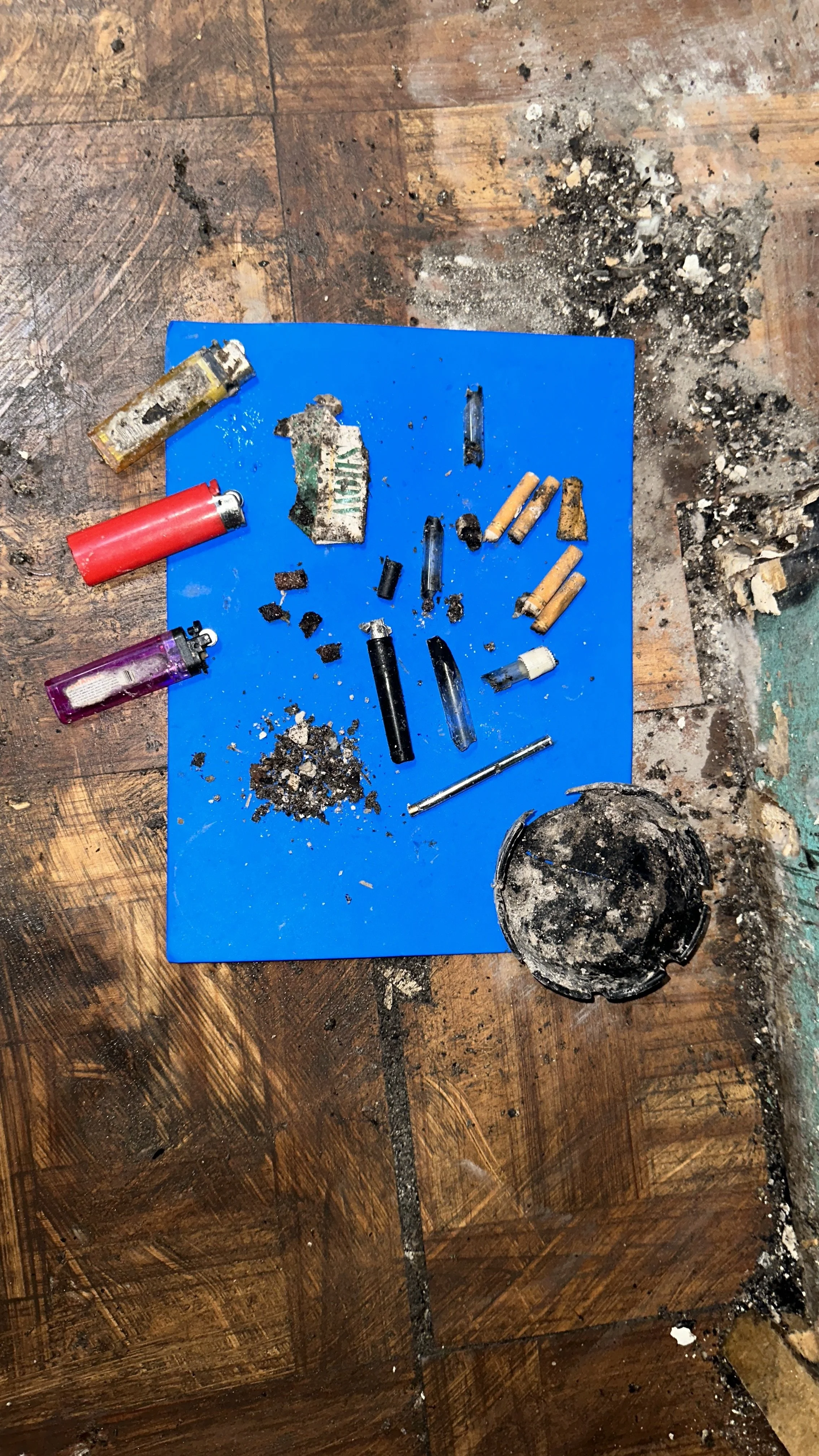 Various burned and unburned cigarette butts, lighters, ash, and debris on a blue surface with a burnt container and ash scattered on a wooden floor.