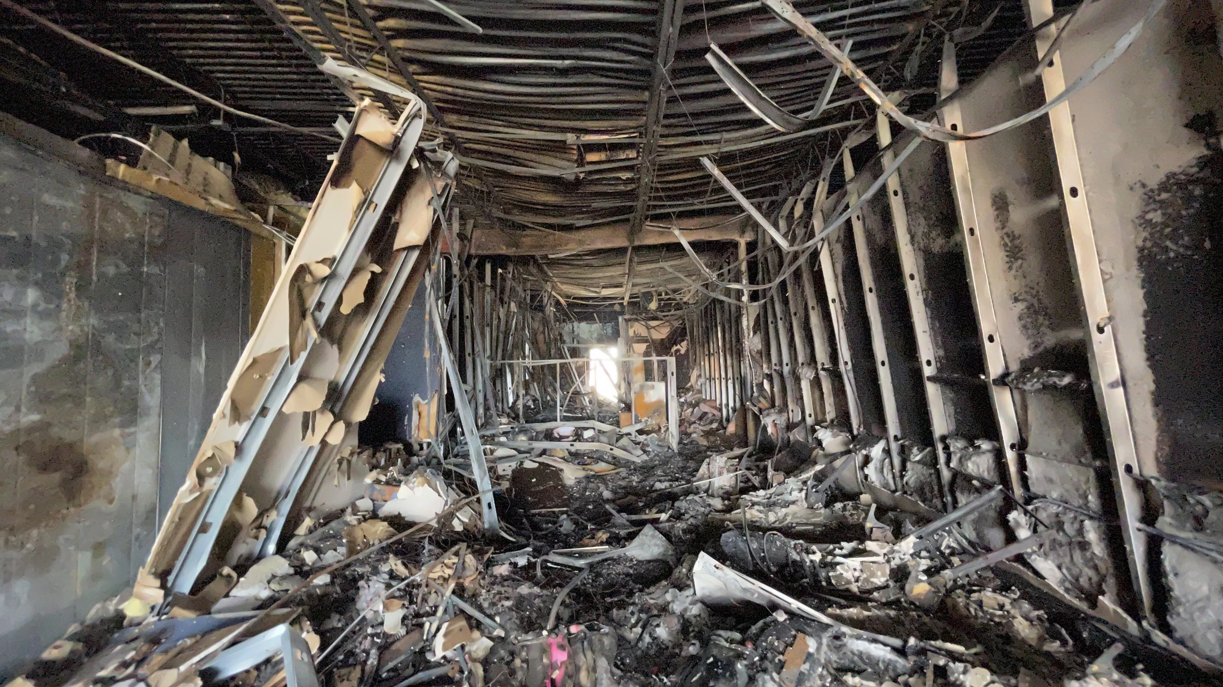 A severely damaged, burned-out corridor with collapsed ceiling, broken wires, and scattered debris.