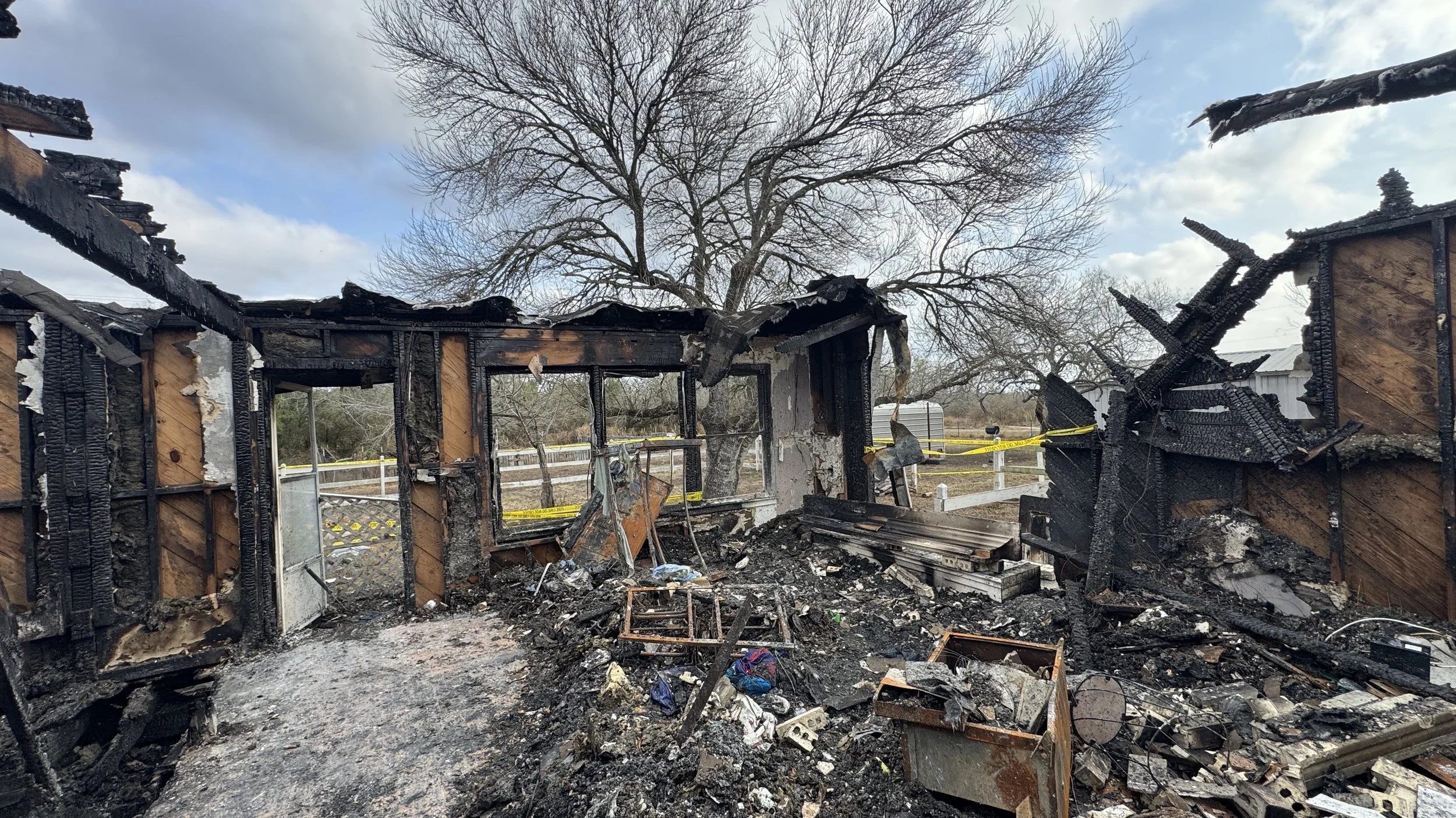 Burned and destroyed building with charred walls and debris, a large tree in the background, and cloudy sky.