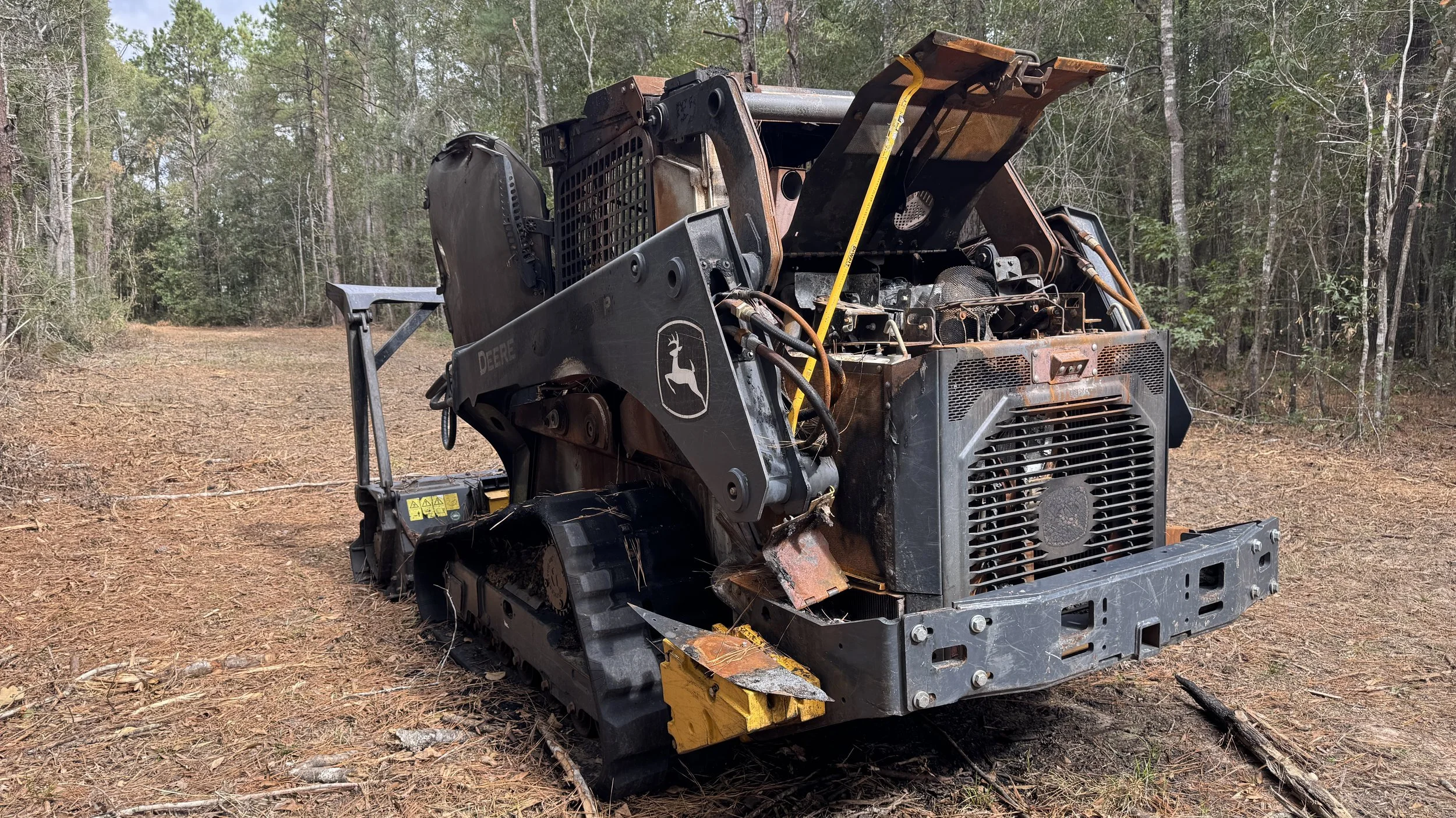 A damaged John Deere skid steer loader with rust and missing parts, sitting on a dirt clearing in a wooded area.