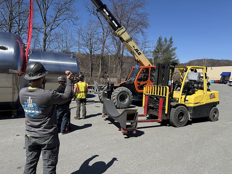 Construction workers and equipment near a large metallic tank, including a yellow forklift and an orange telescopic handler, with some individuals wearing safety gear and helmets, in an outdoor setting.