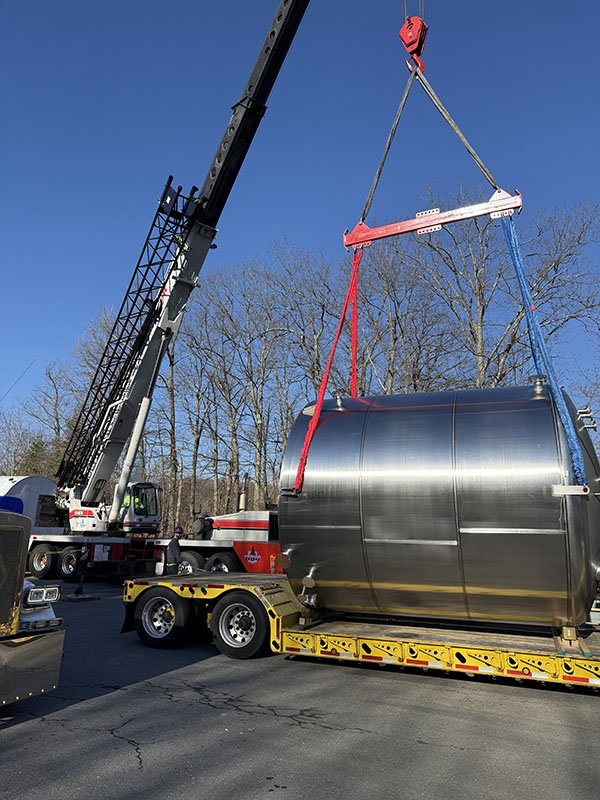 Large industrial crane lifting a shiny metallic cylindrical tank onto a flatbed trailer. The scene takes place outdoors on a clear day, with leafless trees in the background.