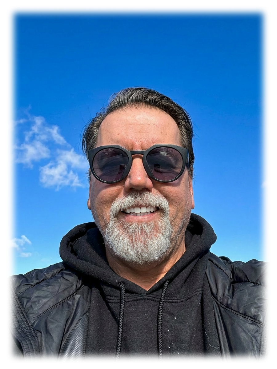 A smiling middle-aged man with a beard and sunglasses, wearing a black hoodie and jacket, outdoors against a bright blue sky with some clouds.