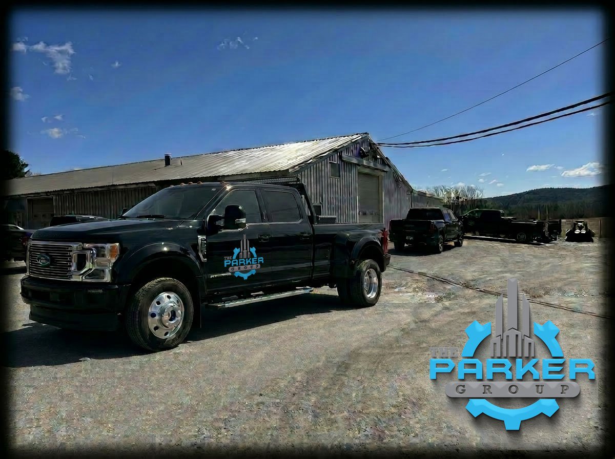 Black pickup truck with The Parker Group logo parked in front of a rustic metal building on a gravel lot, with two other vehicles and a forklift in the background under a blue sky.