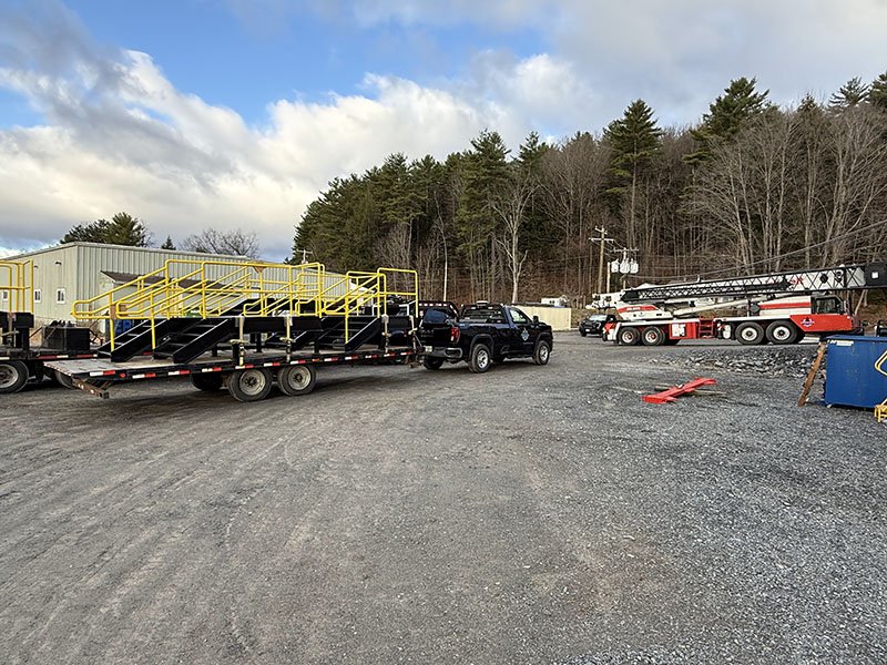 Equipment and vehicles in an outdoor gravel lot, including a trailer with yellow safety railings, multiple black pickup trucks, and a red and white crane truck, with a wooded area and cloudy sky in the background.
