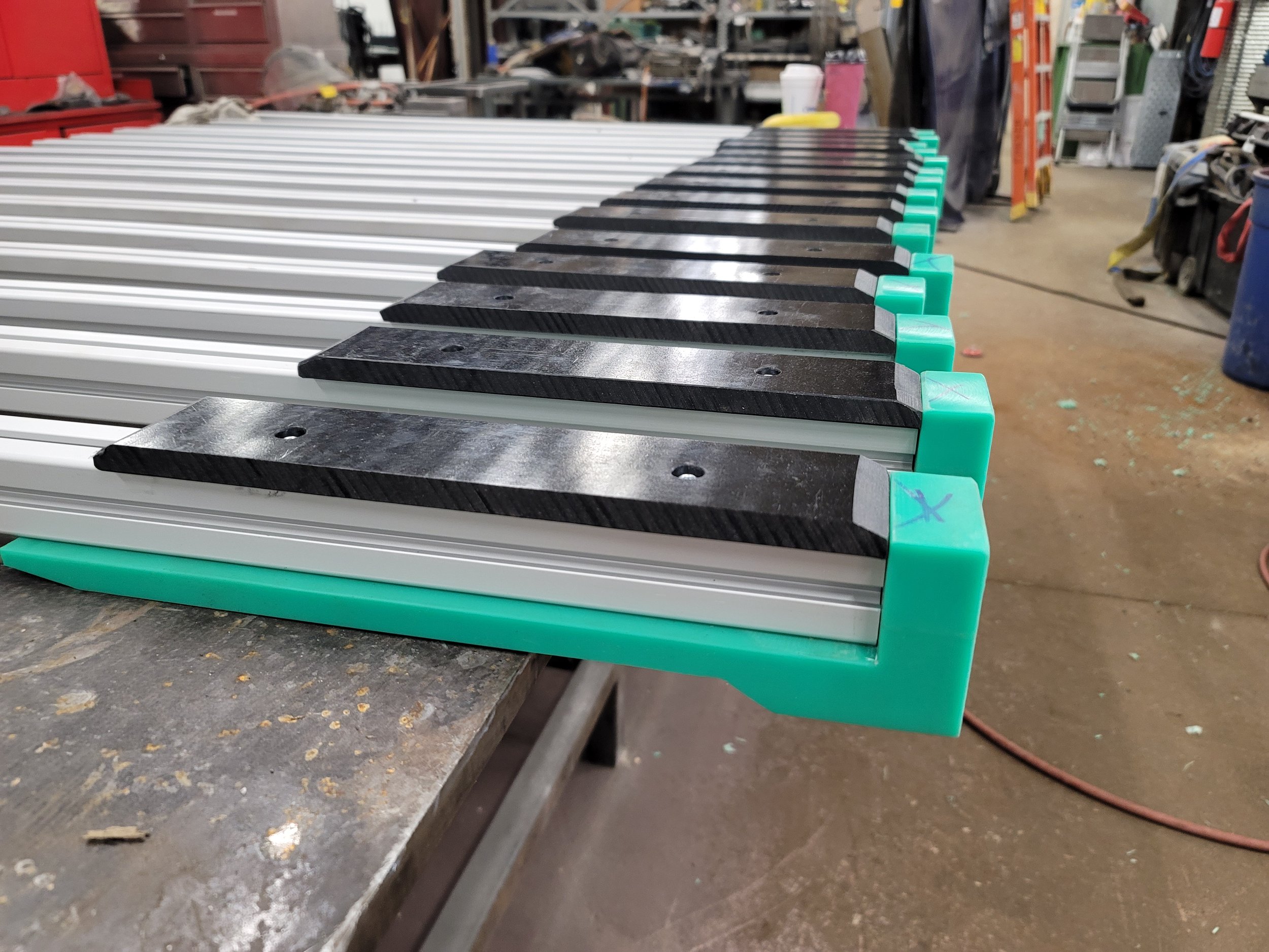 Close-up of a piano keyboard with alternating white and black keys, some with teal-colored finger guards, set in a workshop with tools and equipment in the background.