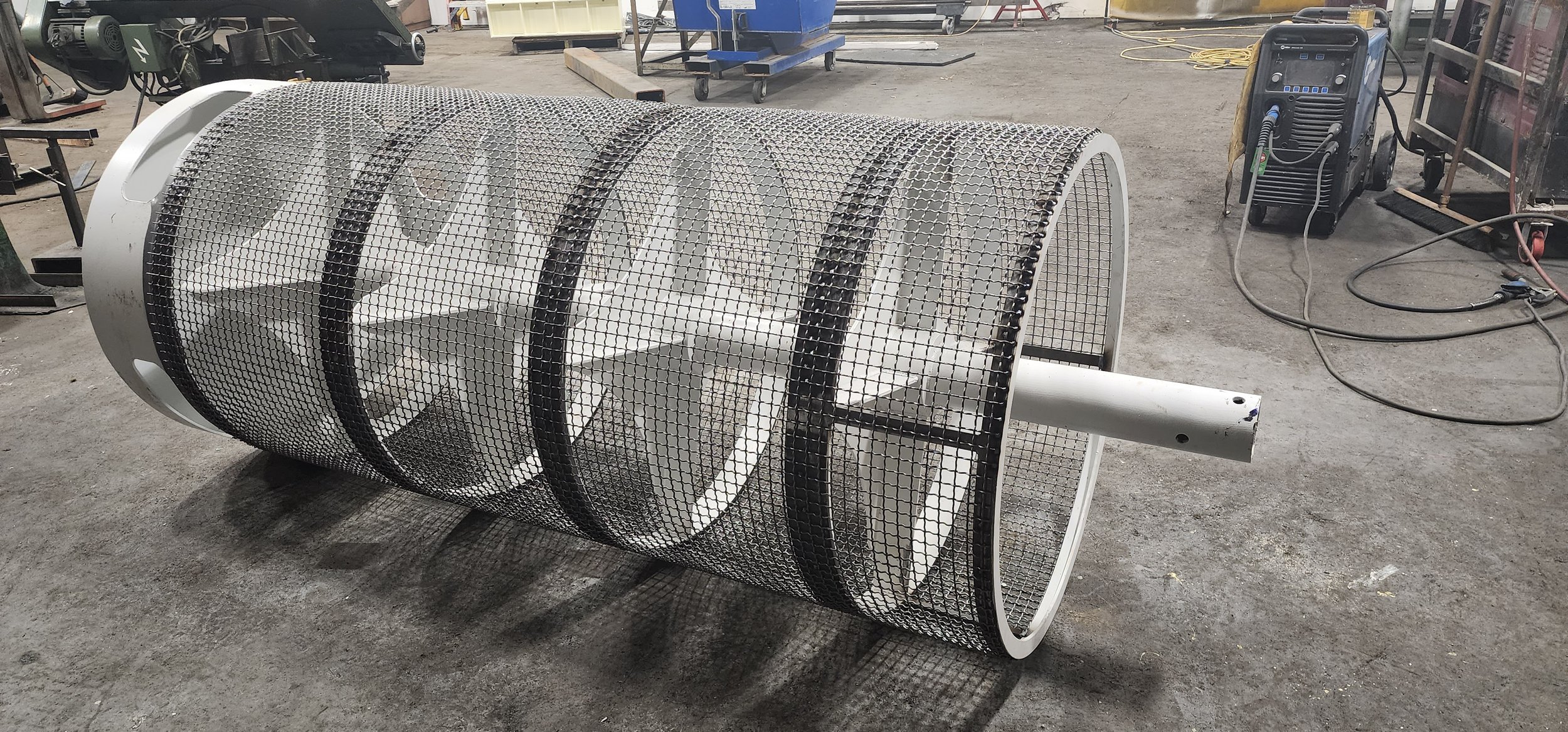 Large cylindrical metal structure with a mesh protective covering on a workshop floor, surrounded by industrial equipment and tools.