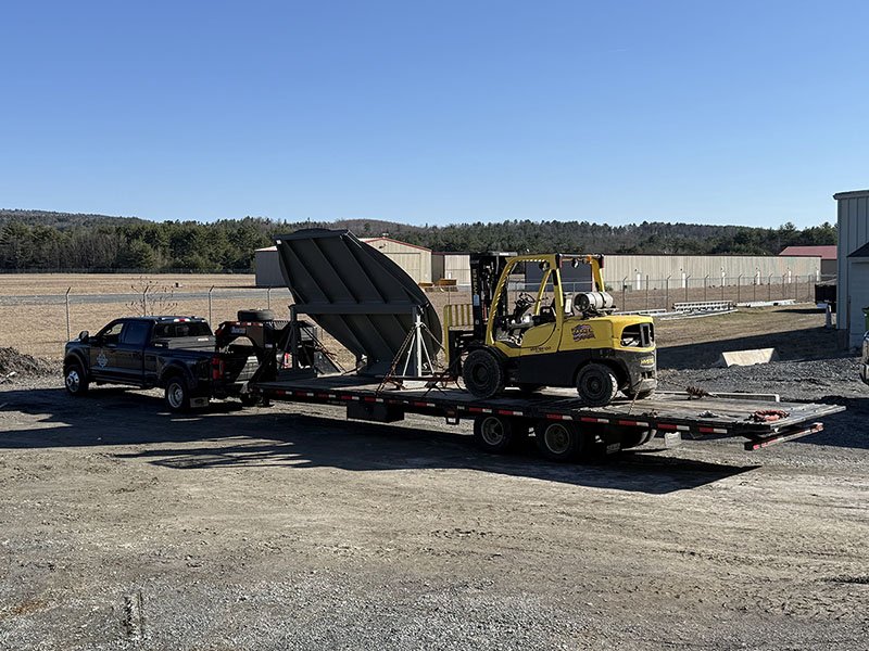 A flatbed trailer transporting a yellow forklift and a large black metal structure in an industrial area with warehouses, dirt ground, and a fence in the background under a clear blue sky.