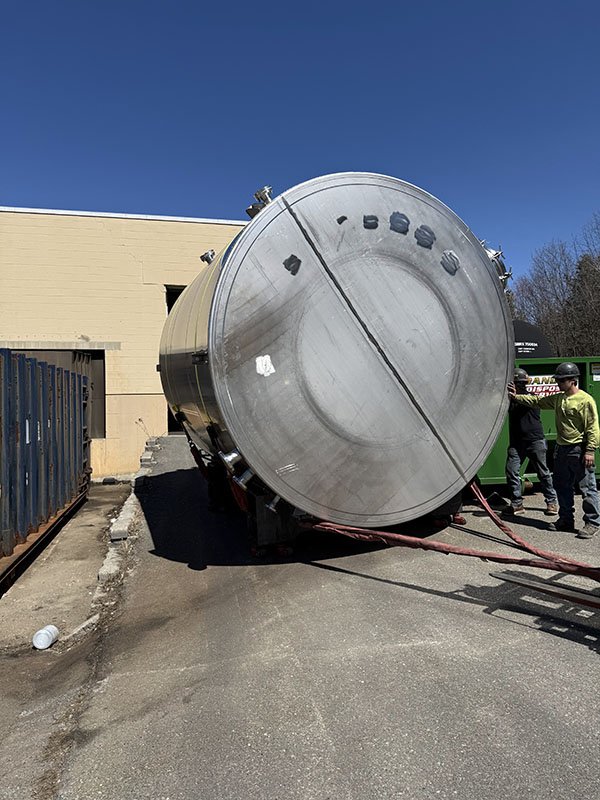 Large cylindrical metallic tank lying on its side outdoors, with a bright blue sky and a beige building in the background. Several people are nearby, some wearing helmets.