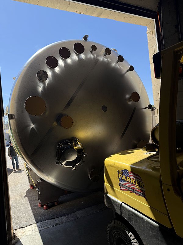 Large cylindrical metal tank or vessel outside a building, with a blue sky and some people in the background, and a yellow construction vehicle with a logo that reads "The Parker Group".