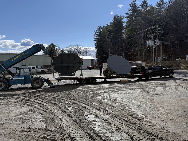 A flatbed truck being loaded with large satellite dishes by a blue forklift, with a few people nearby, on a dirt lot under a blue sky with some clouds and trees in the background.