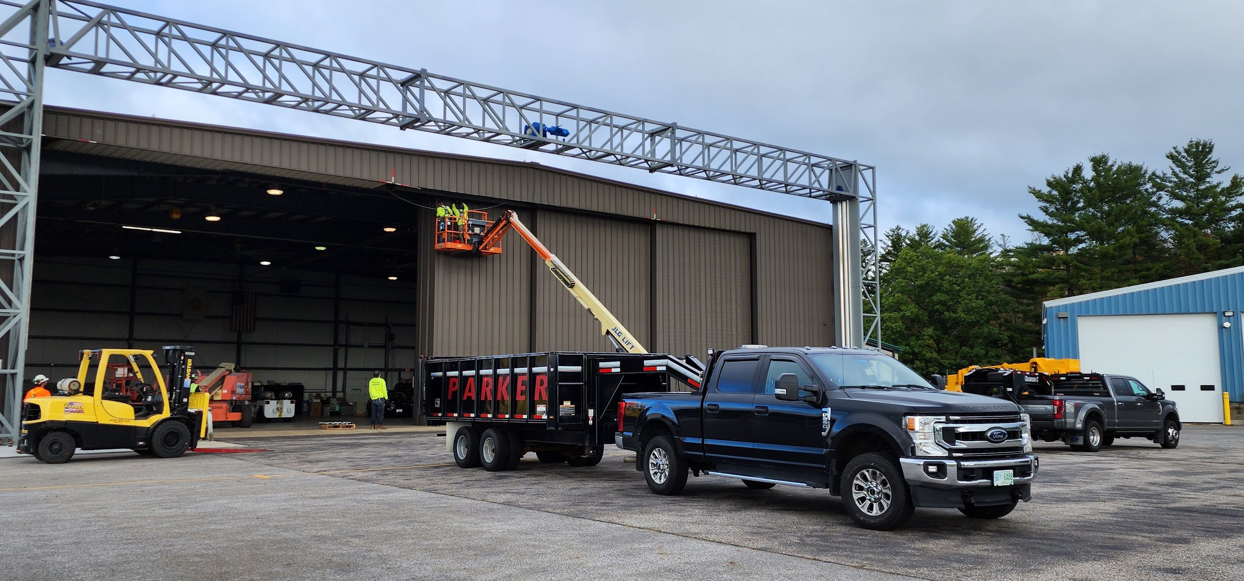 Workers installing a metal structure on a large industrial building with various vehicles including a forklift, trucks, and an extension lift.
