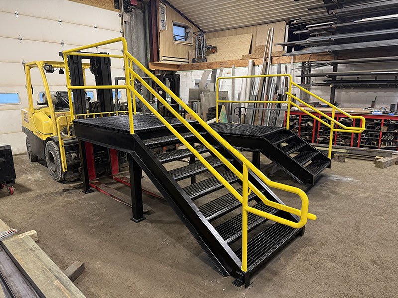 A black metal industrial staircase with yellow handrails in a workshop, connected to a yellow forklift.