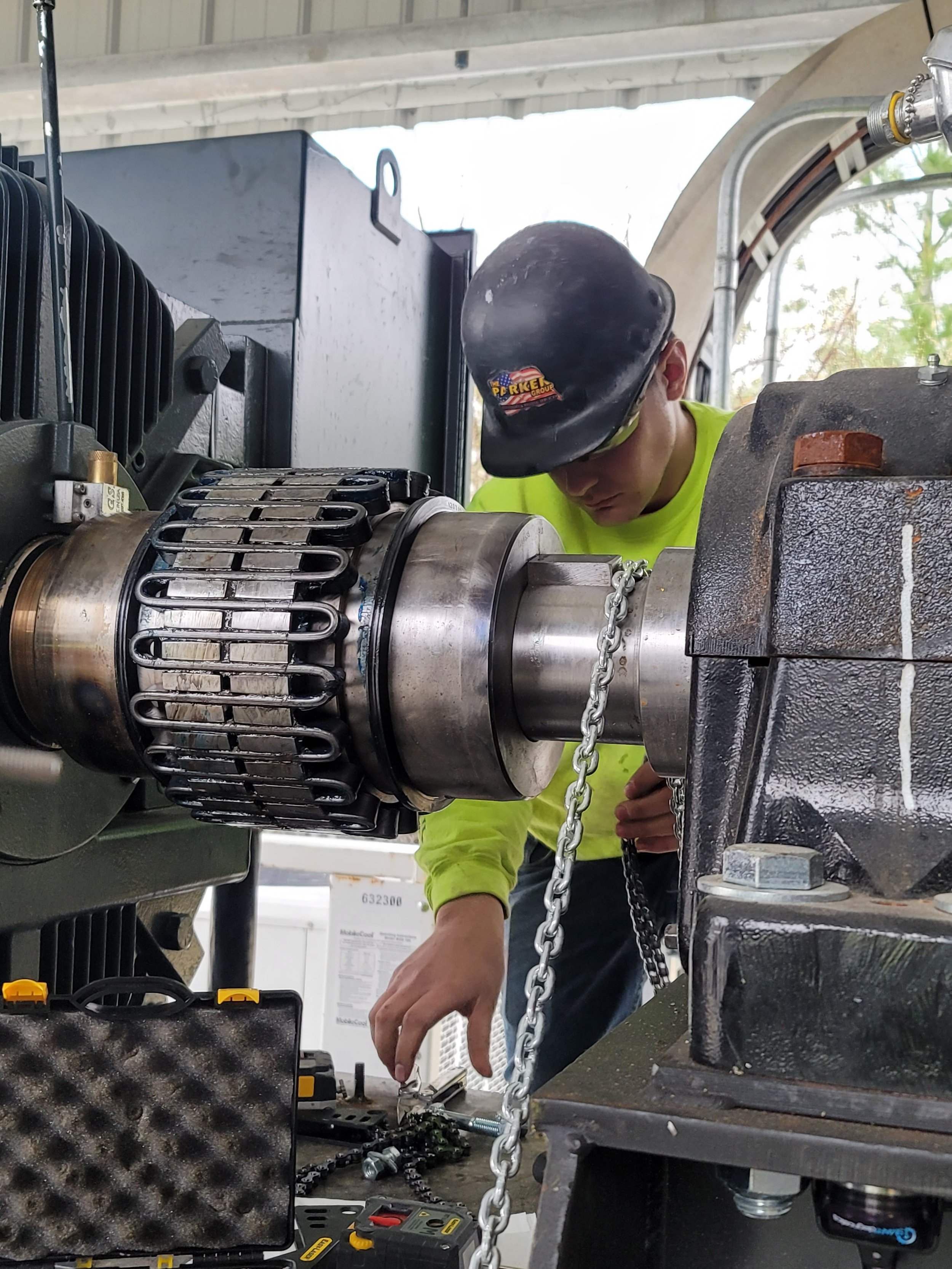 A worker wearing a safety helmet and neon yellow shirt is working on a large industrial machine or engine, using tools on the machinery in a workshop or industrial setting.