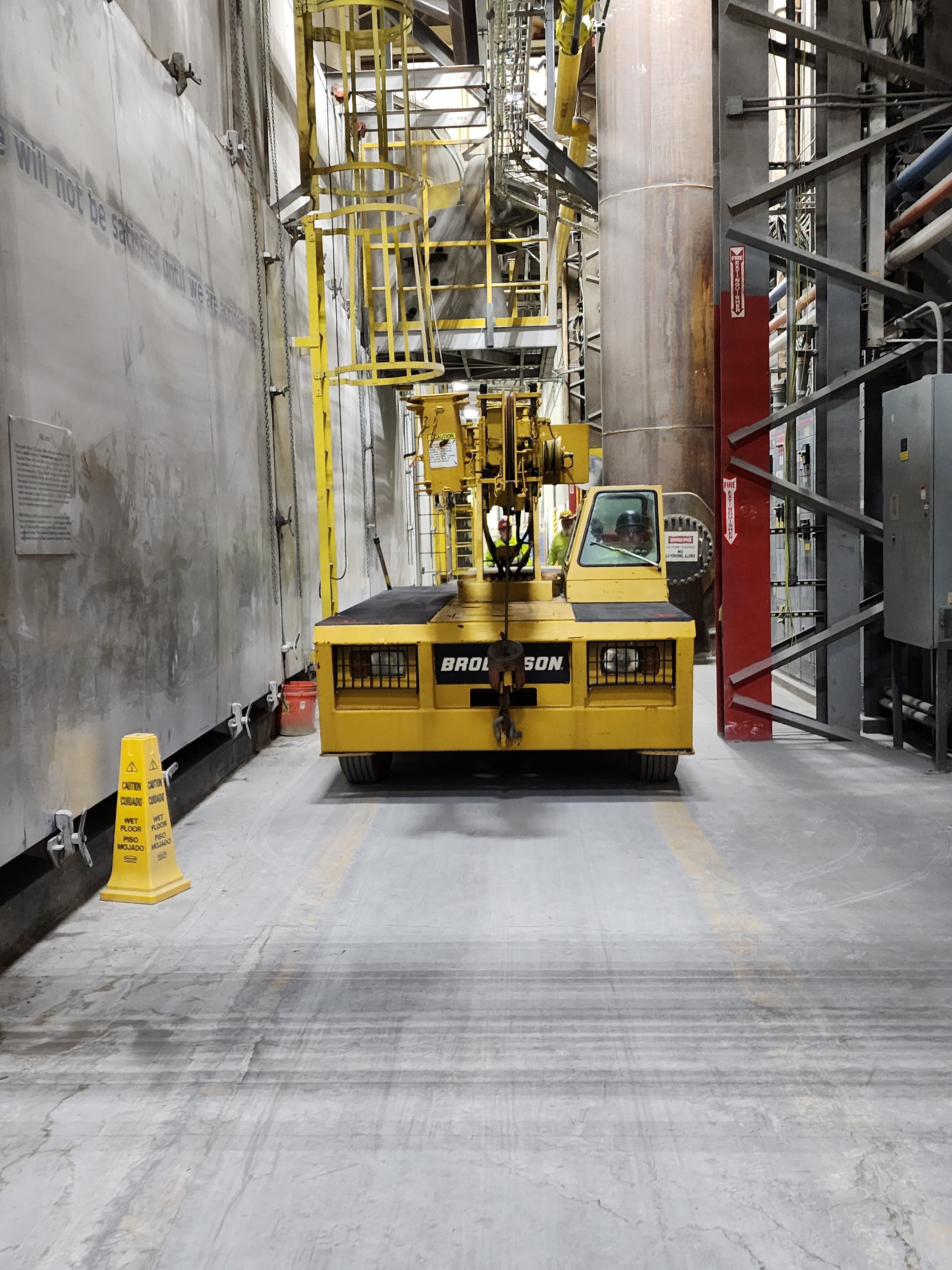 Industrial setting with a yellow forklift in the center, surrounded by safety barriers, pipes, and equipment. Yellow caution signs are on the ground, and workers in safety gear are visible in the background.