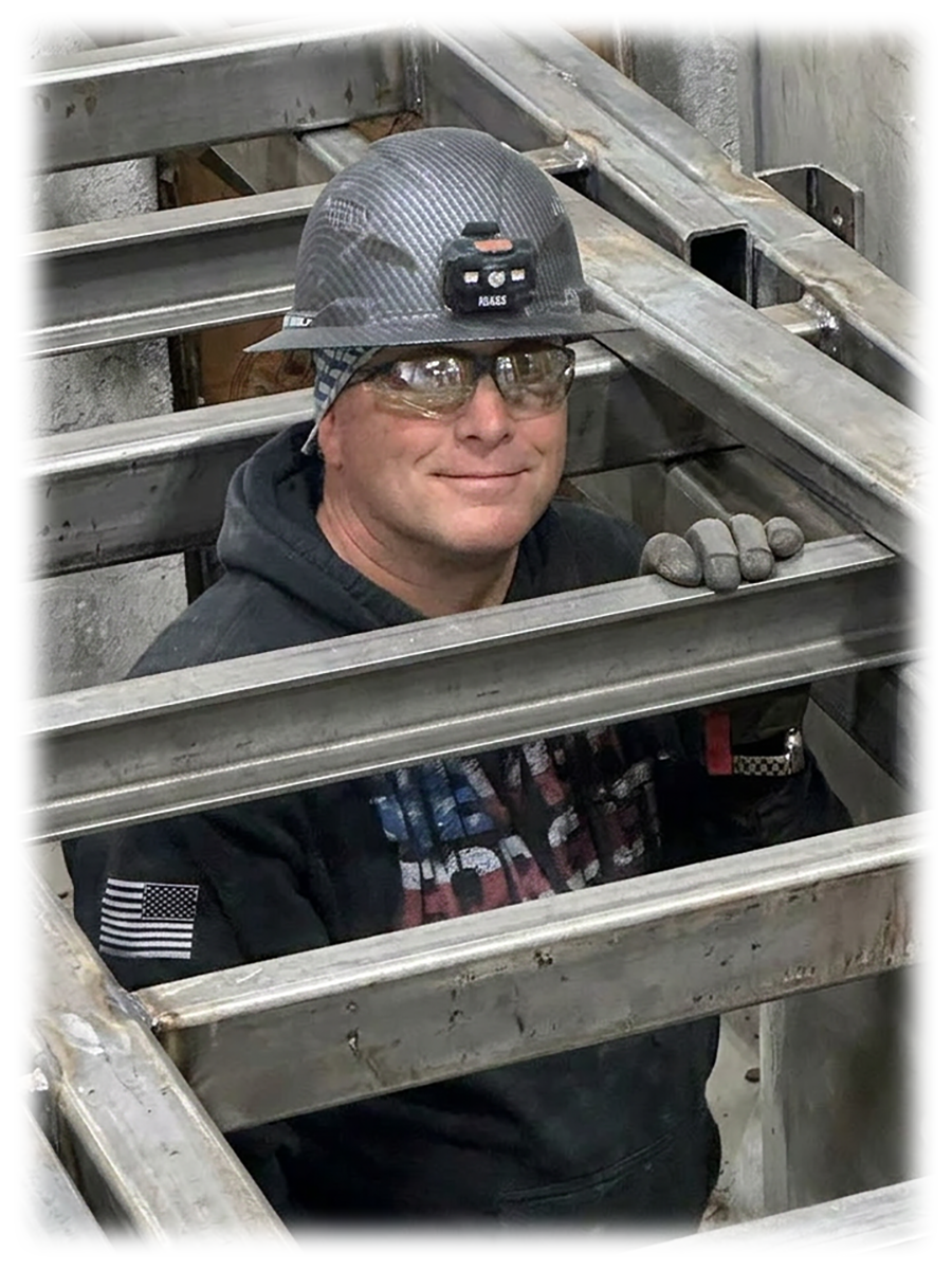 A construction worker wearing a carbon fiber helmet with a headlamp, safety glasses, gloves, and a hoodie, smiling while working among metal beams in a building or workshop.